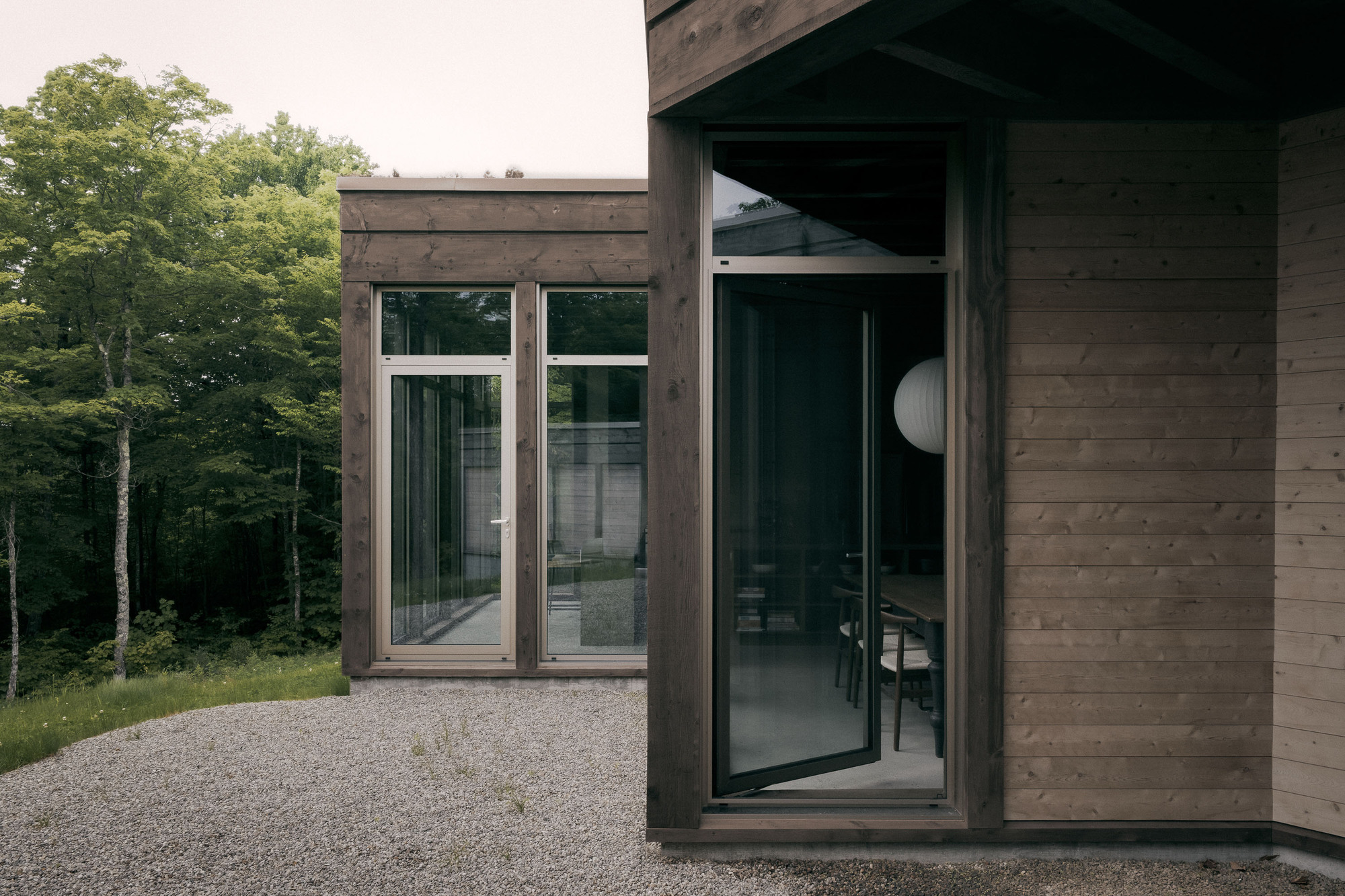Corner of two timber-clad volumes with glazed doors, opening onto a gravel court beside a stand of birch trees