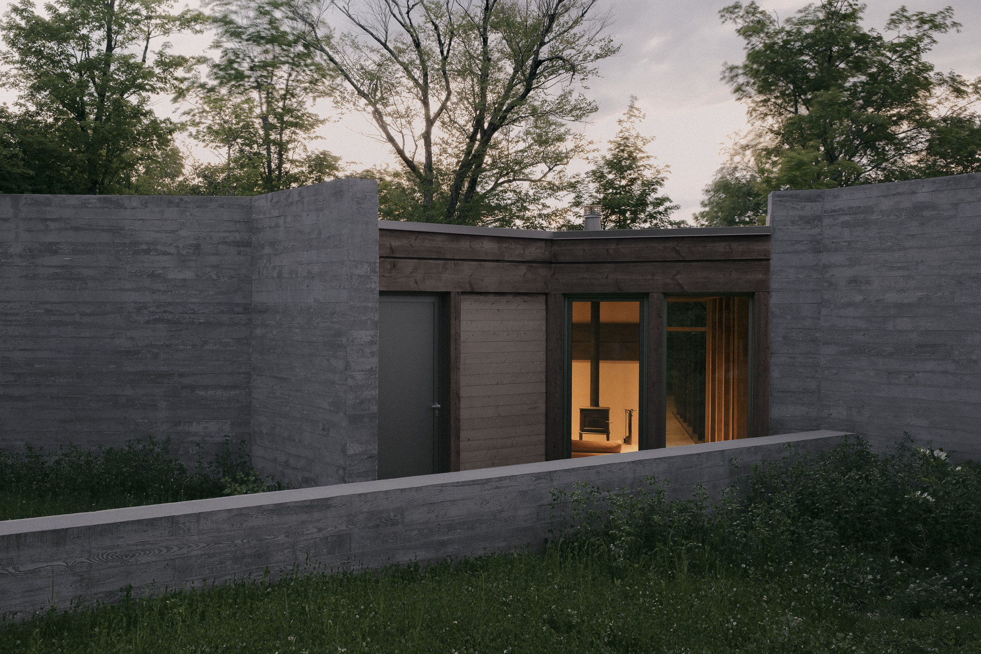 Entry court framed by board-formed concrete walls, with a lit fireplace room glowing through glass at twilight