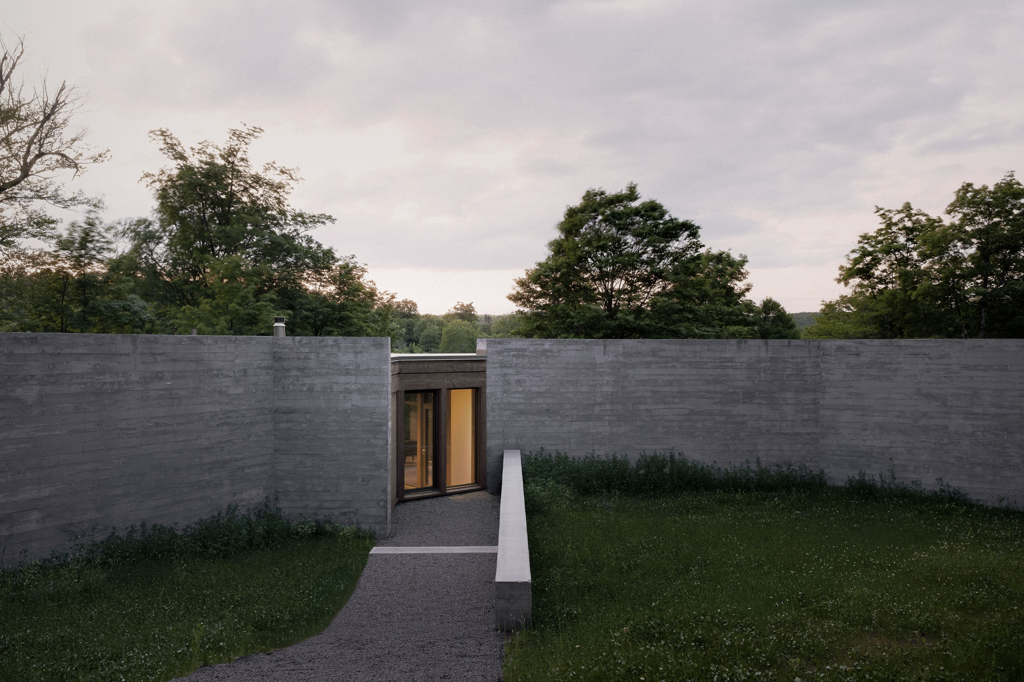 Board-formed concrete walls flanking a gravel path that leads to a glazed timber entrance, under an overcast dusk sky