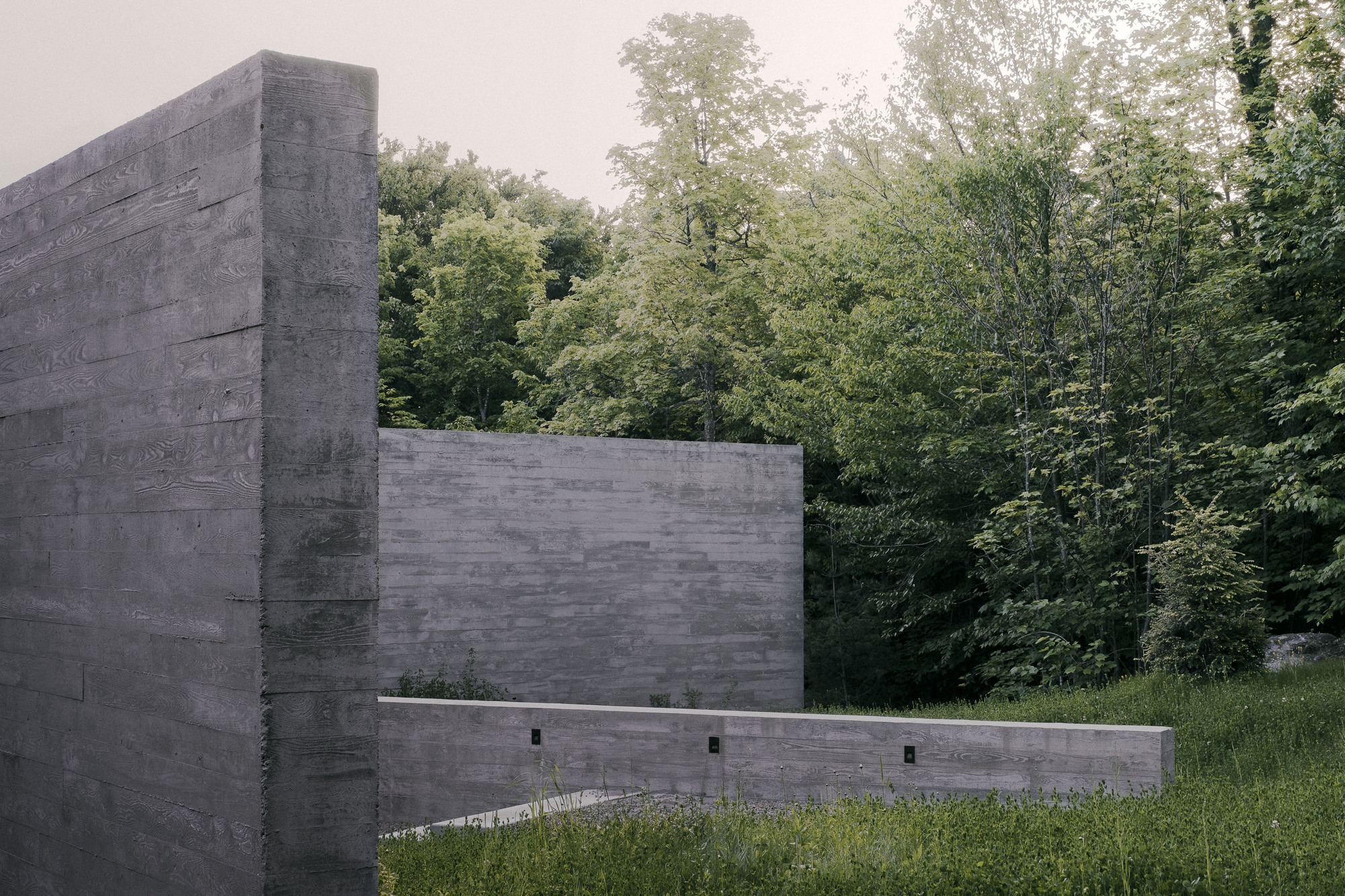 Board-formed concrete wall corner and low retaining wall with small lights, set against dense green forest