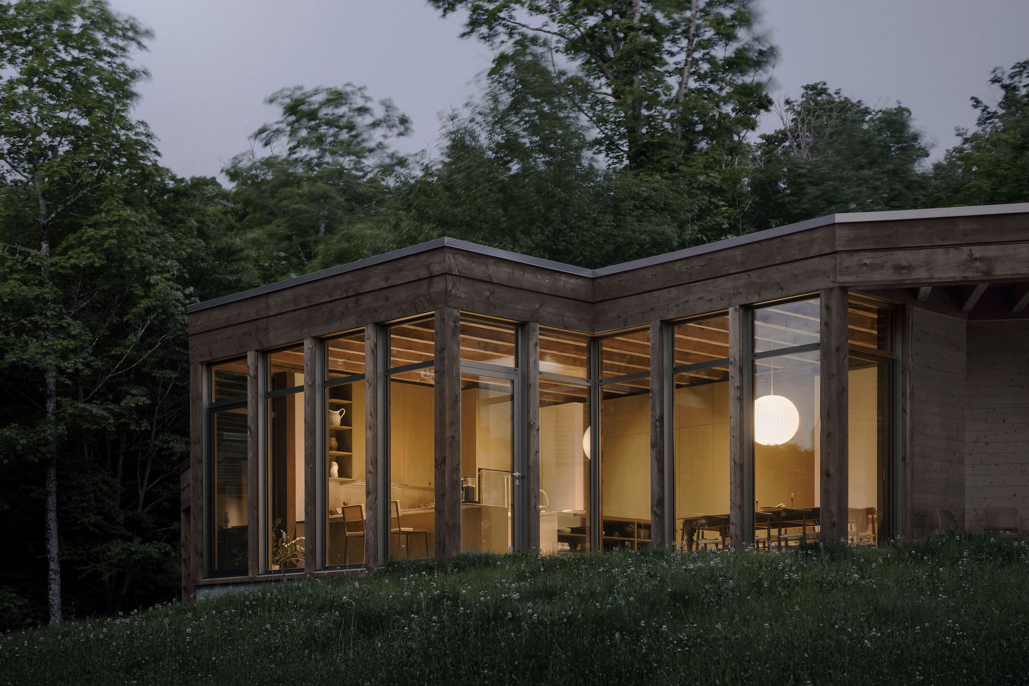 Lit interior with a round pendant lamp and dining table glowing through floor-to-ceiling glazing at nightfall