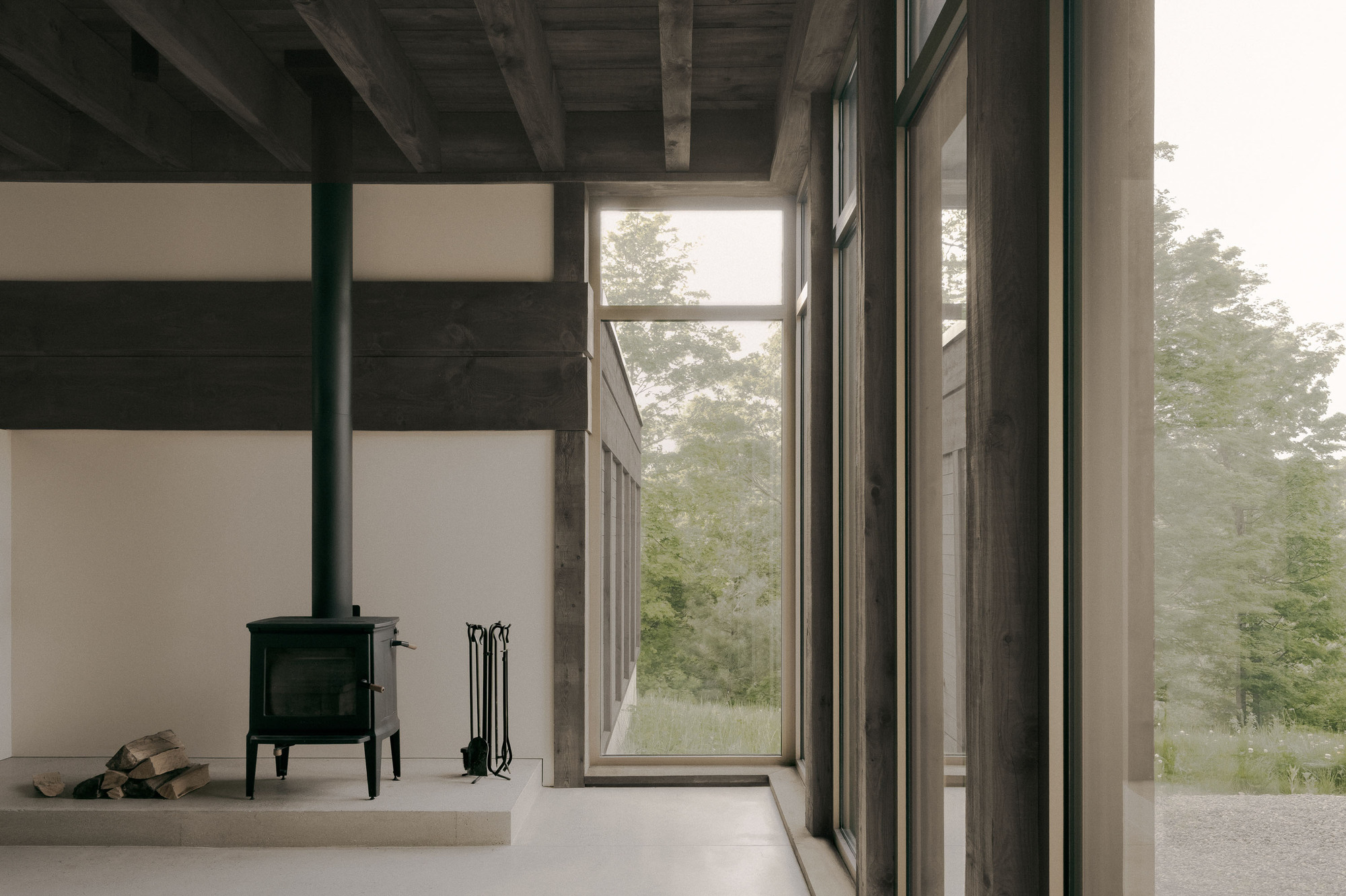Black wood stove on a raised concrete hearth with stacked logs, framed by weathered timber posts and tall windows onto misty trees