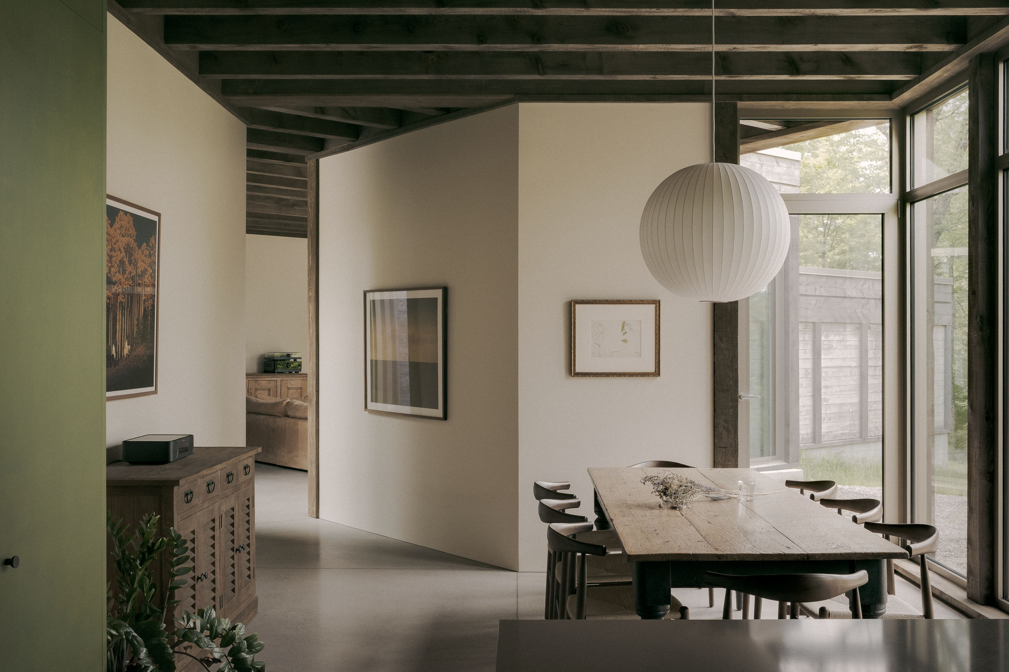 Dining area with a long wooden table, elbow chairs and a round paper pendant lamp beneath exposed timber joists