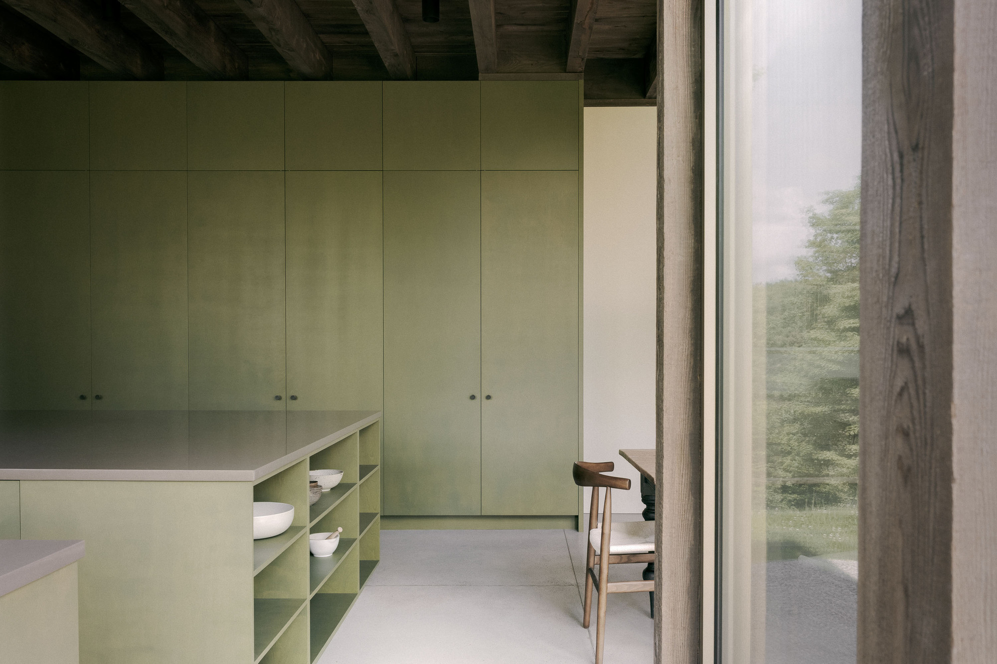 Olive green kitchen with full-height cabinets and an open-shelf island, a timber post and glazing in the foreground
