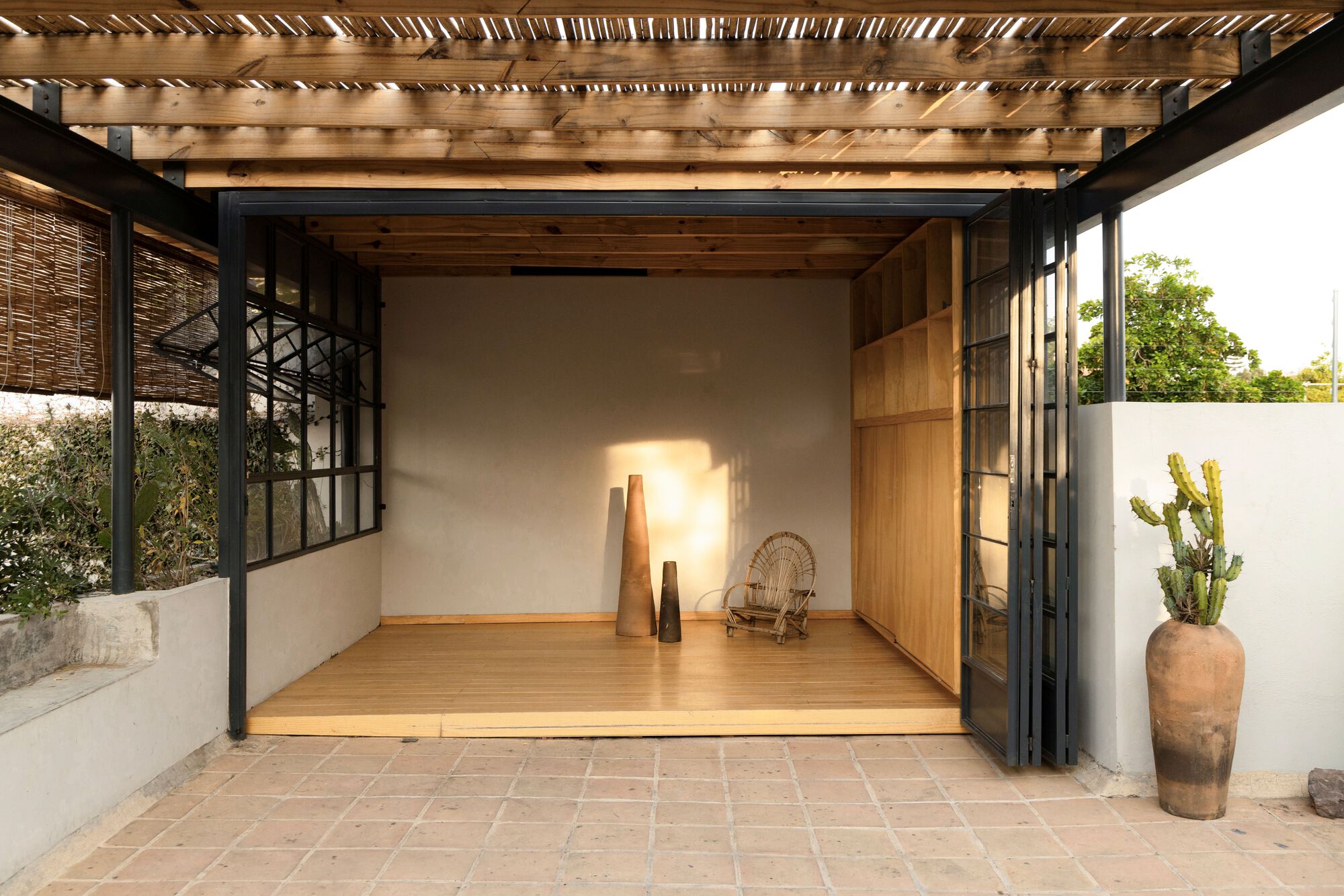 Open rooftop room with timber floor and plywood cabinetry framed by black steel folding doors, a potted cactus beside it in late light