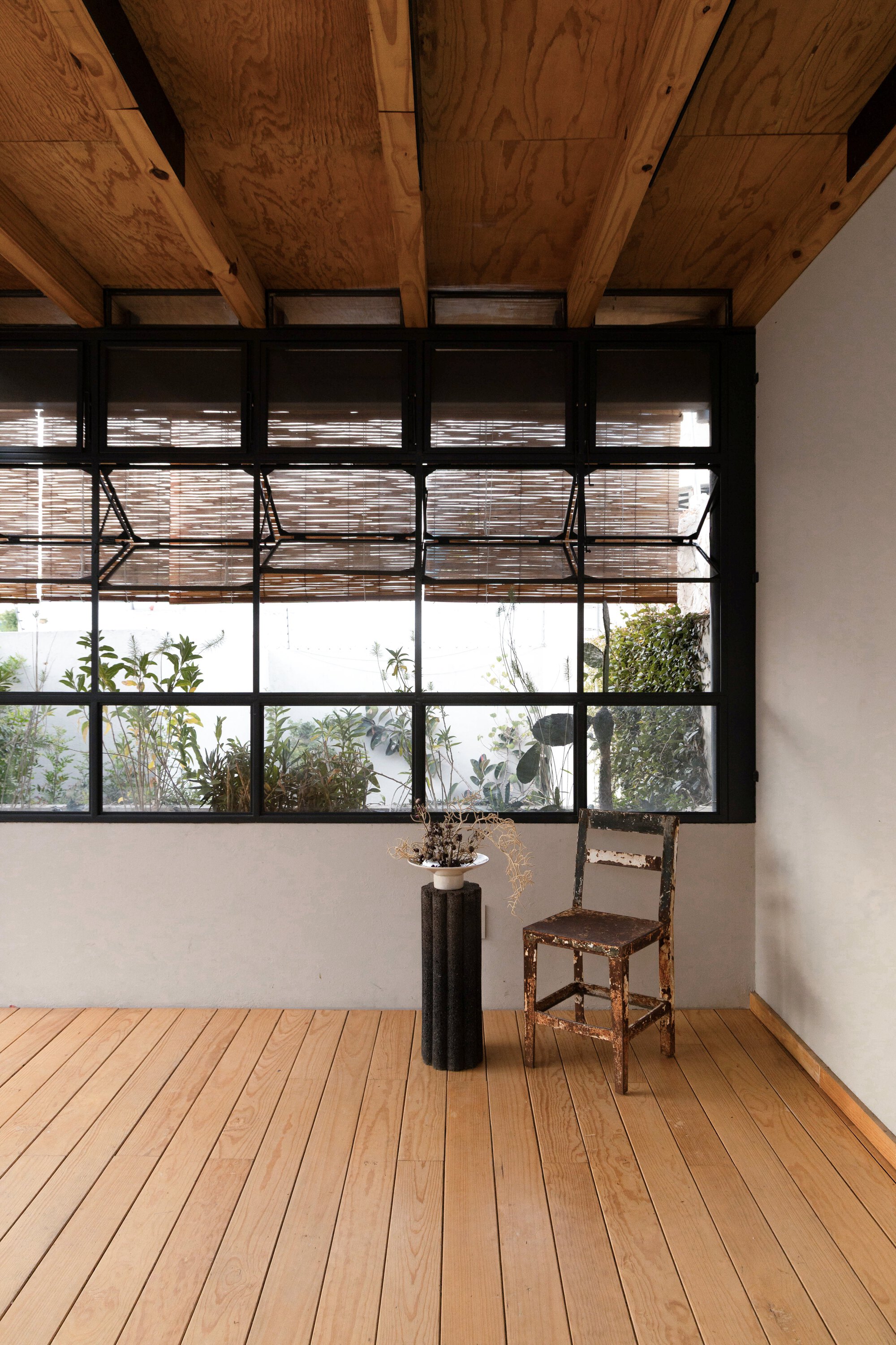 Interior corner with steel-framed hopper windows, exposed timber beams and a worn wooden chair beside a dark column vase on a plank floor