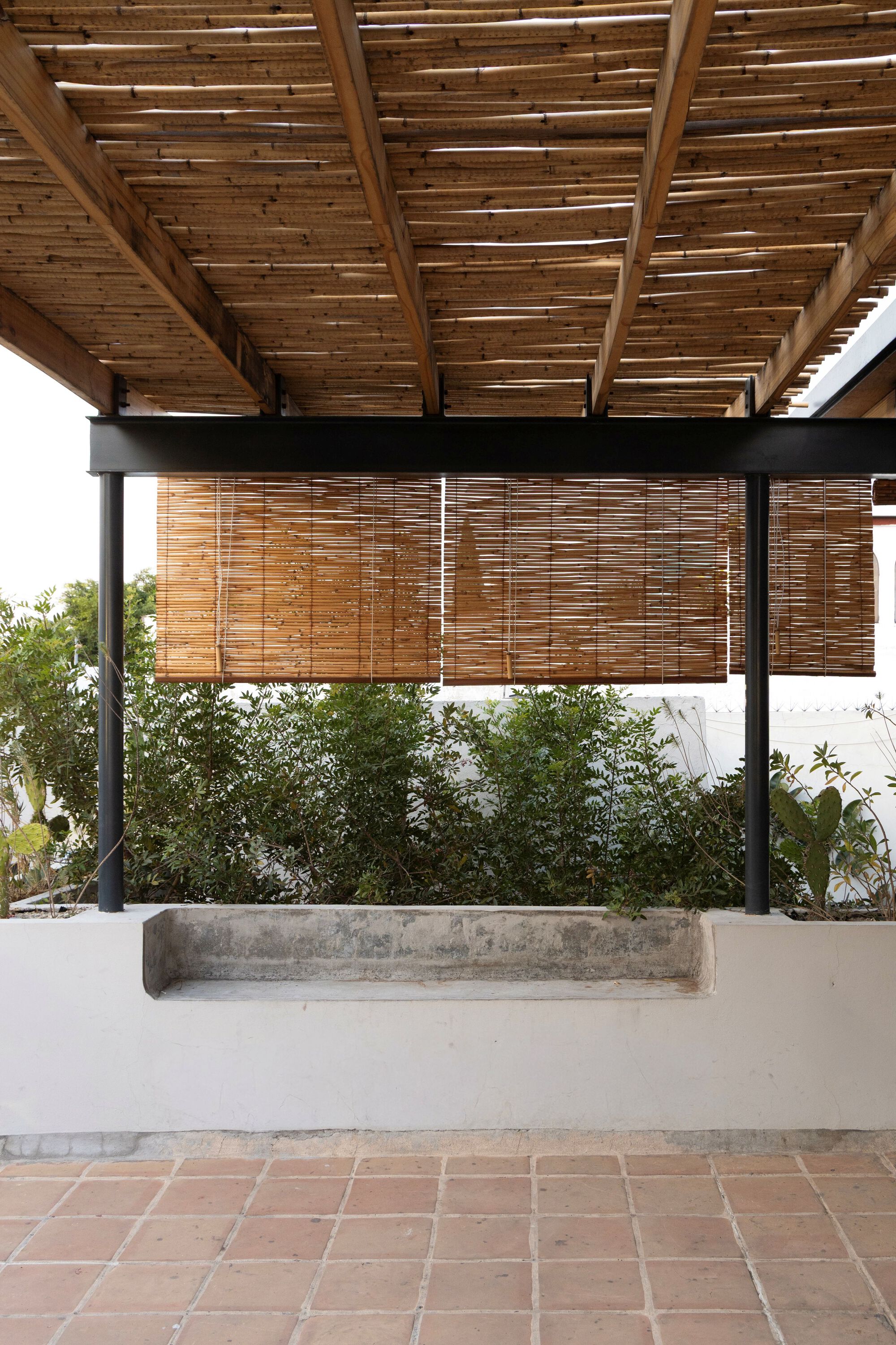 Covered terrace with steel frame, cane ceiling and lowered bamboo blinds above a concrete planter bench and terracotta tile floor