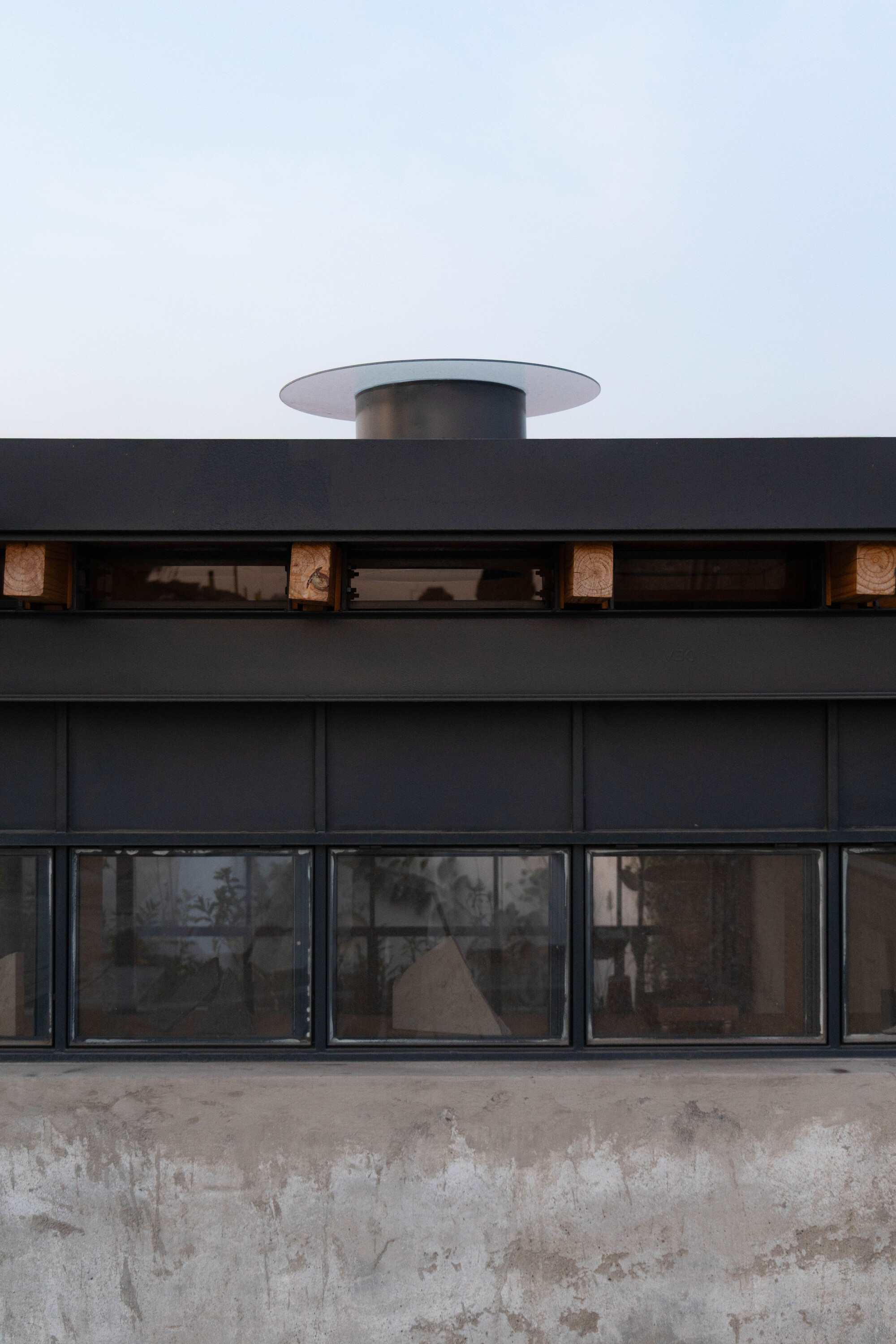 Black steel clerestory band with timber blocks and a round roof vent above a weathered concrete wall under pale sky