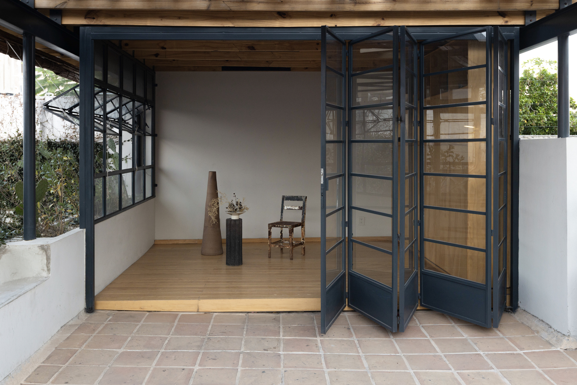 Folding steel-framed glass doors opened onto a timber platform with a tall clay vessel and a single chair, terracotta tiles in front