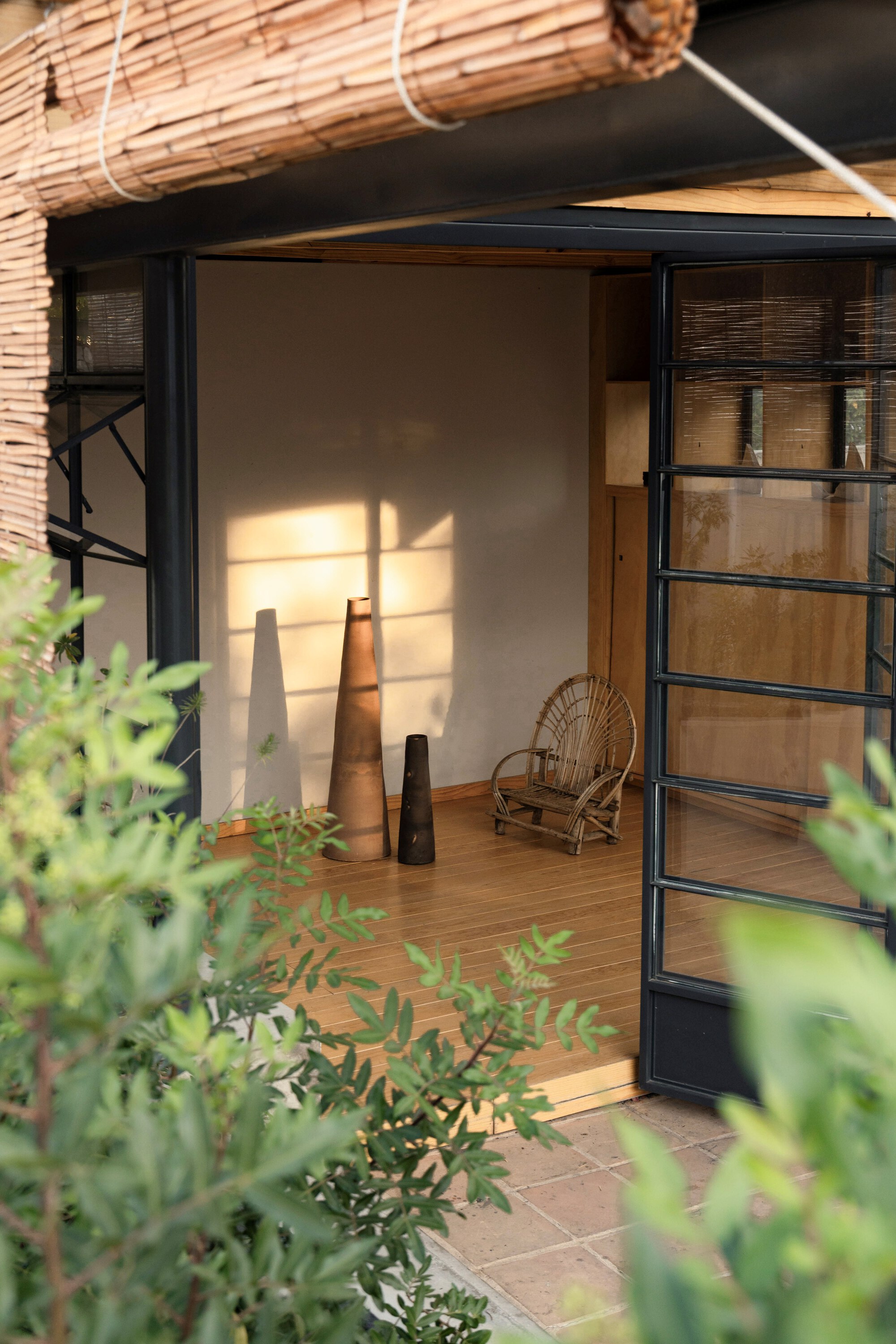 Raised bamboo blind over a steel-framed opening, with late sun falling across a plaster wall, clay vessels and a wicker chair