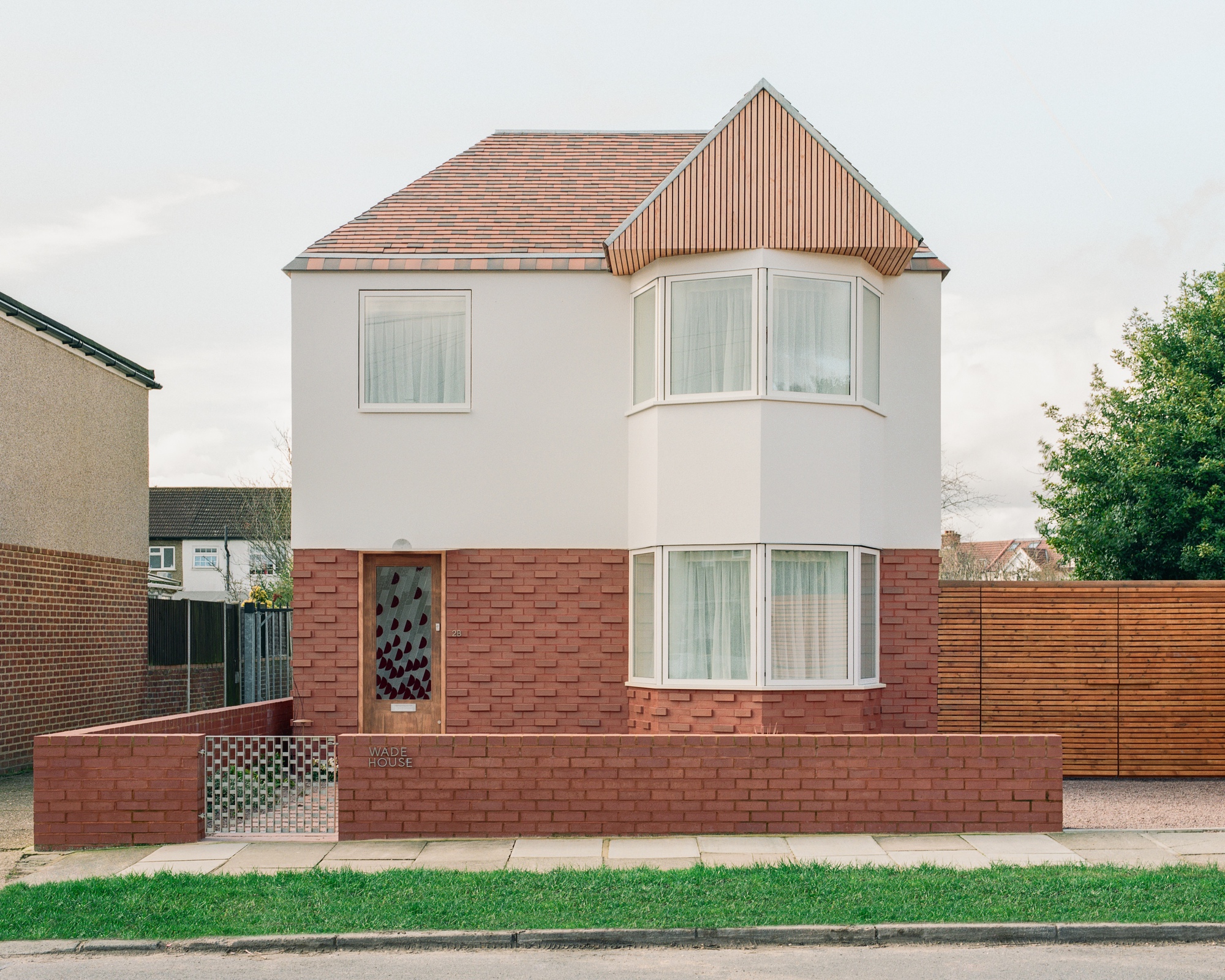 Front facade of a white rendered house with red brick base, bay window and timber-clad gable under an overcast sky