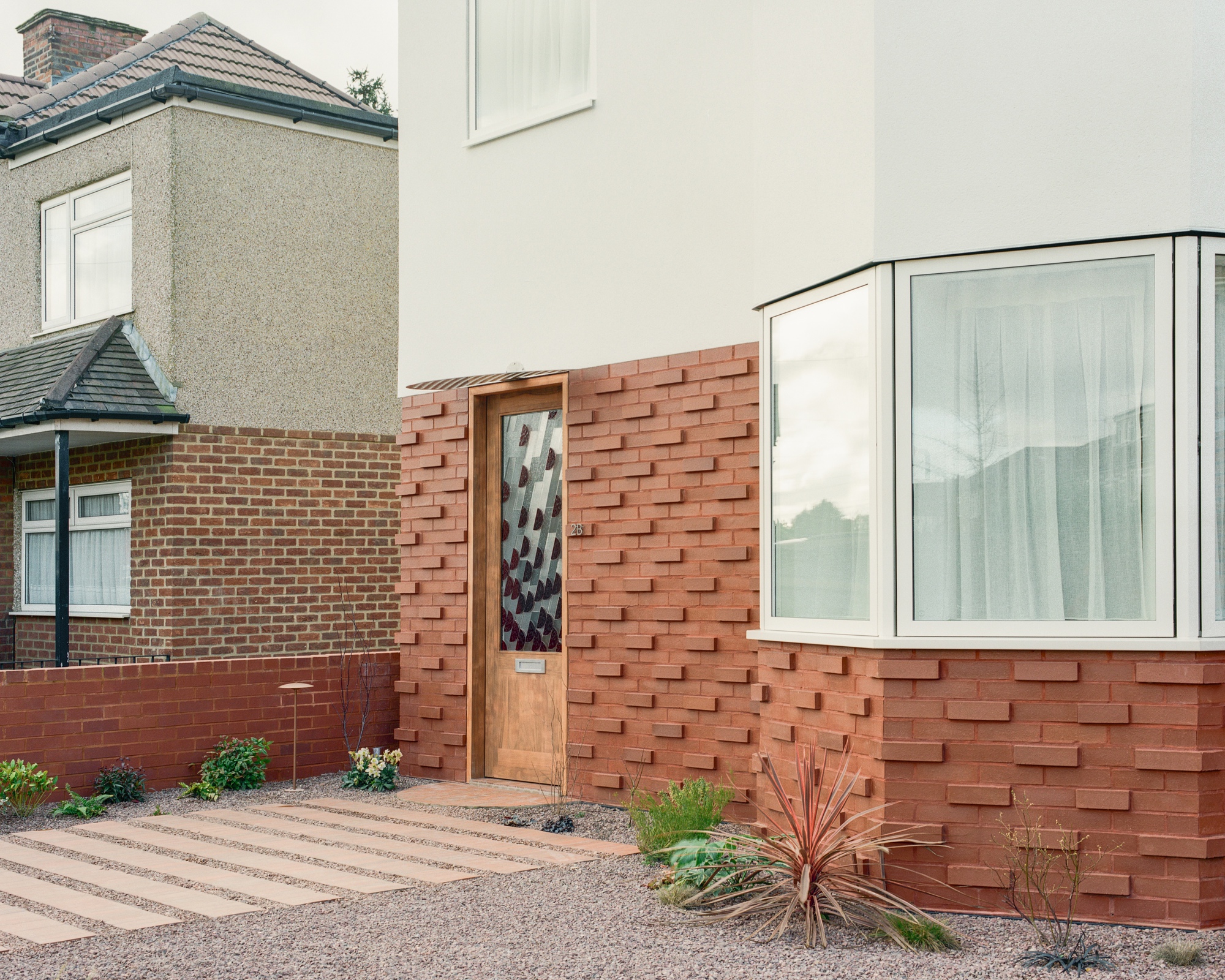 Timber front door set in a textured red brick wall beside a bay window, with gravel and spiky plants in front