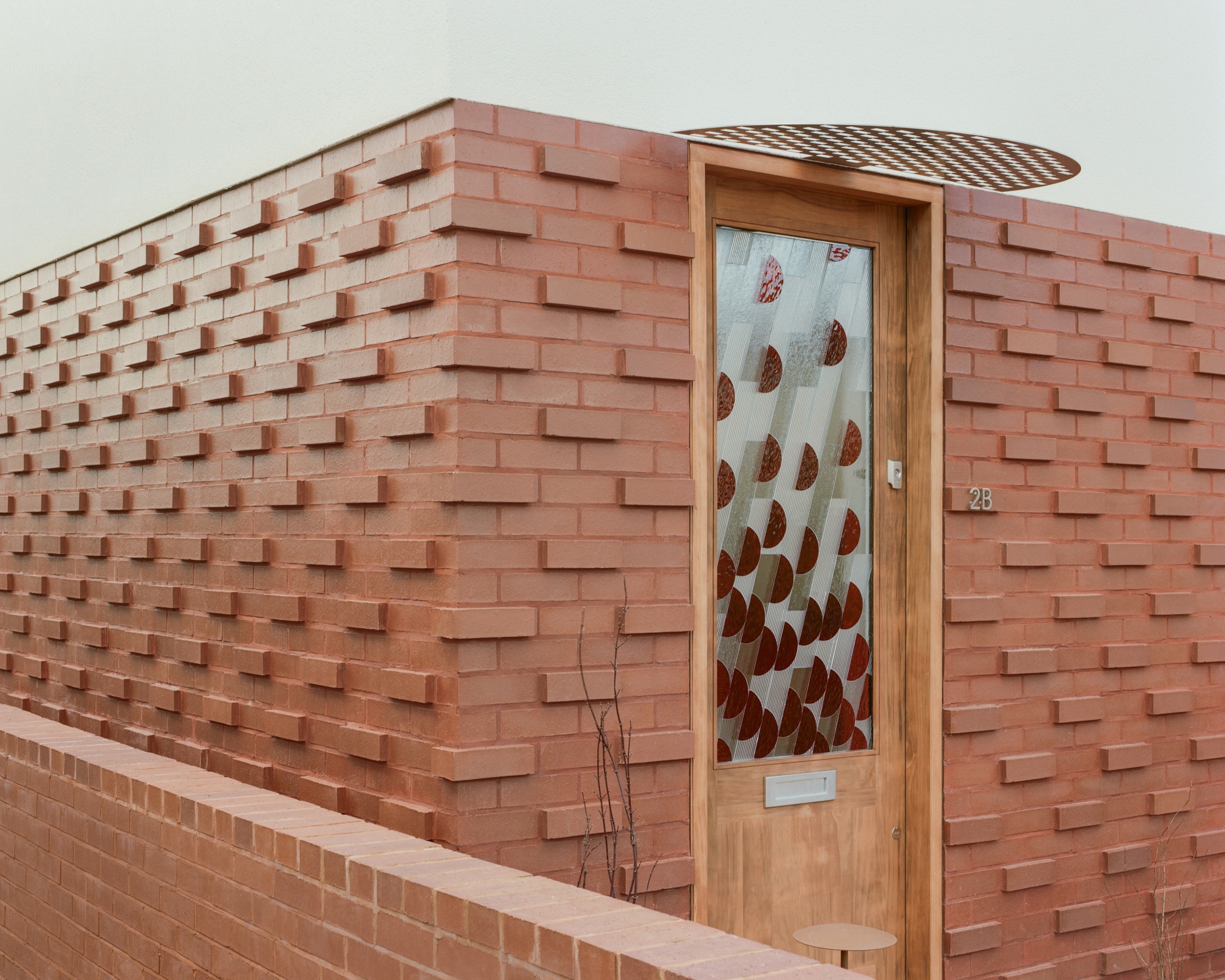 Corner of projecting brick wall with a timber-framed door, patterned glass panel and perforated canopy in soft daylight