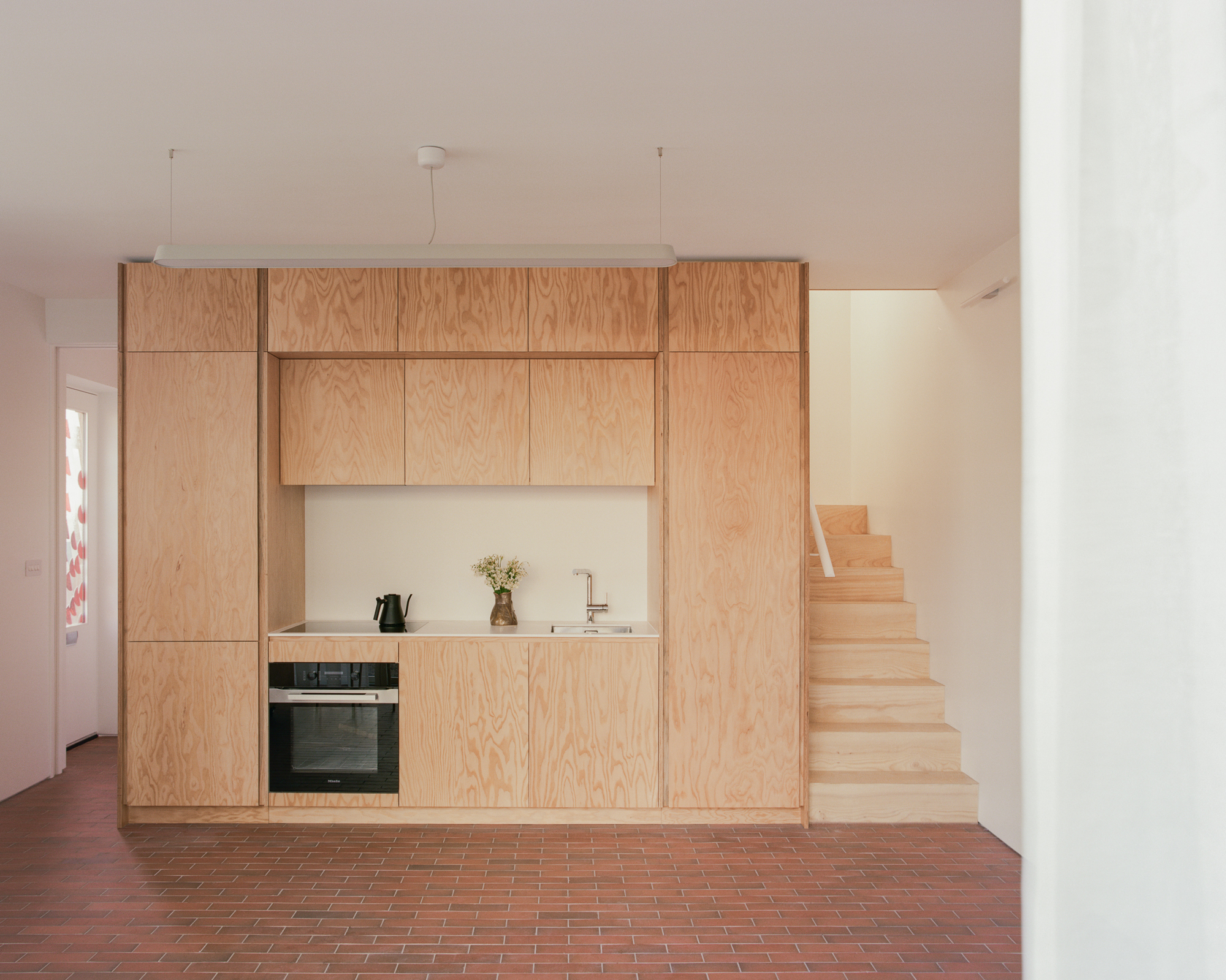 Plywood kitchen block with built-in oven and sink beside a timber stair, on a red tile floor