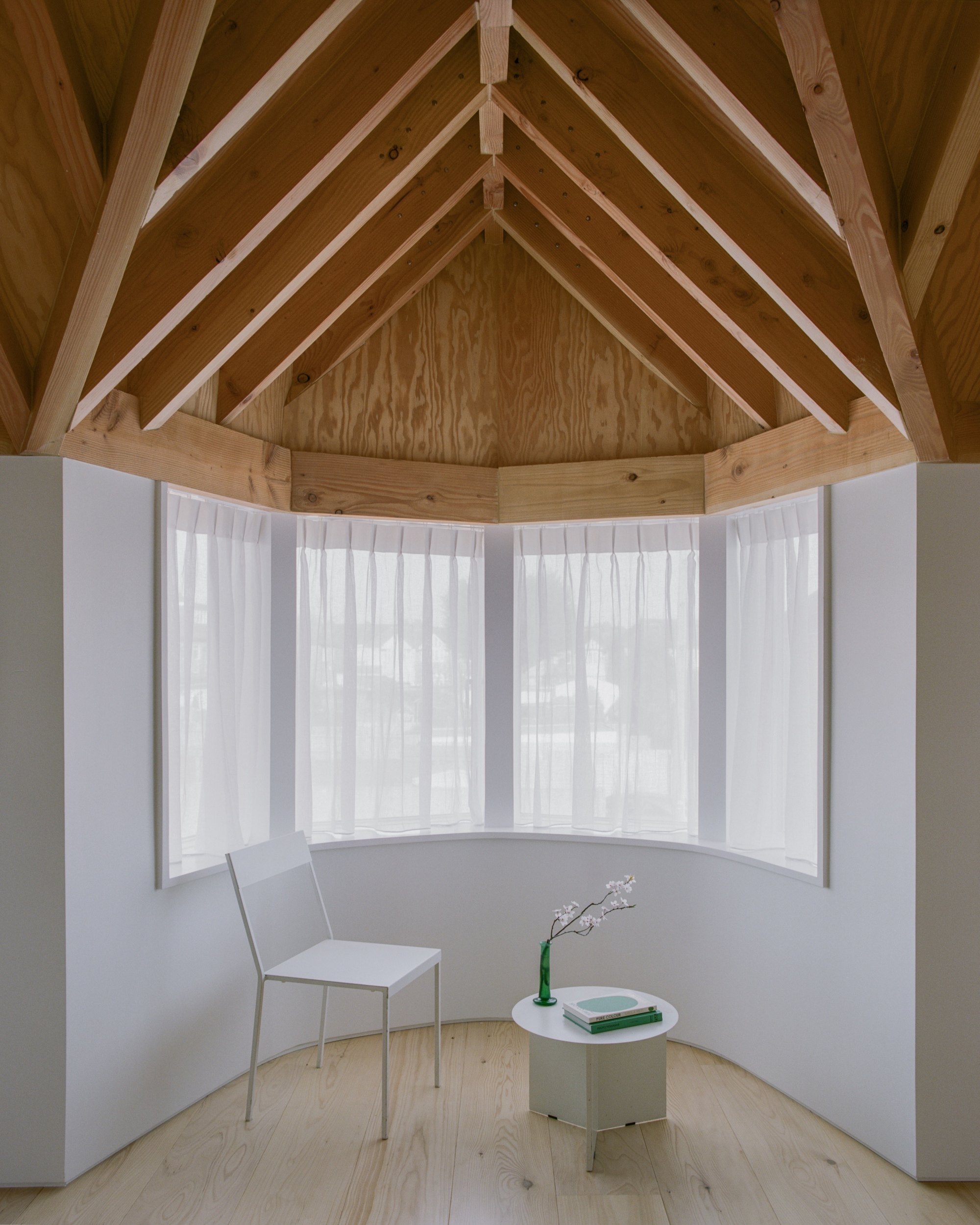 Curved bay window alcove with exposed timber rafters, sheer curtains, a white chair and small side table in daylight