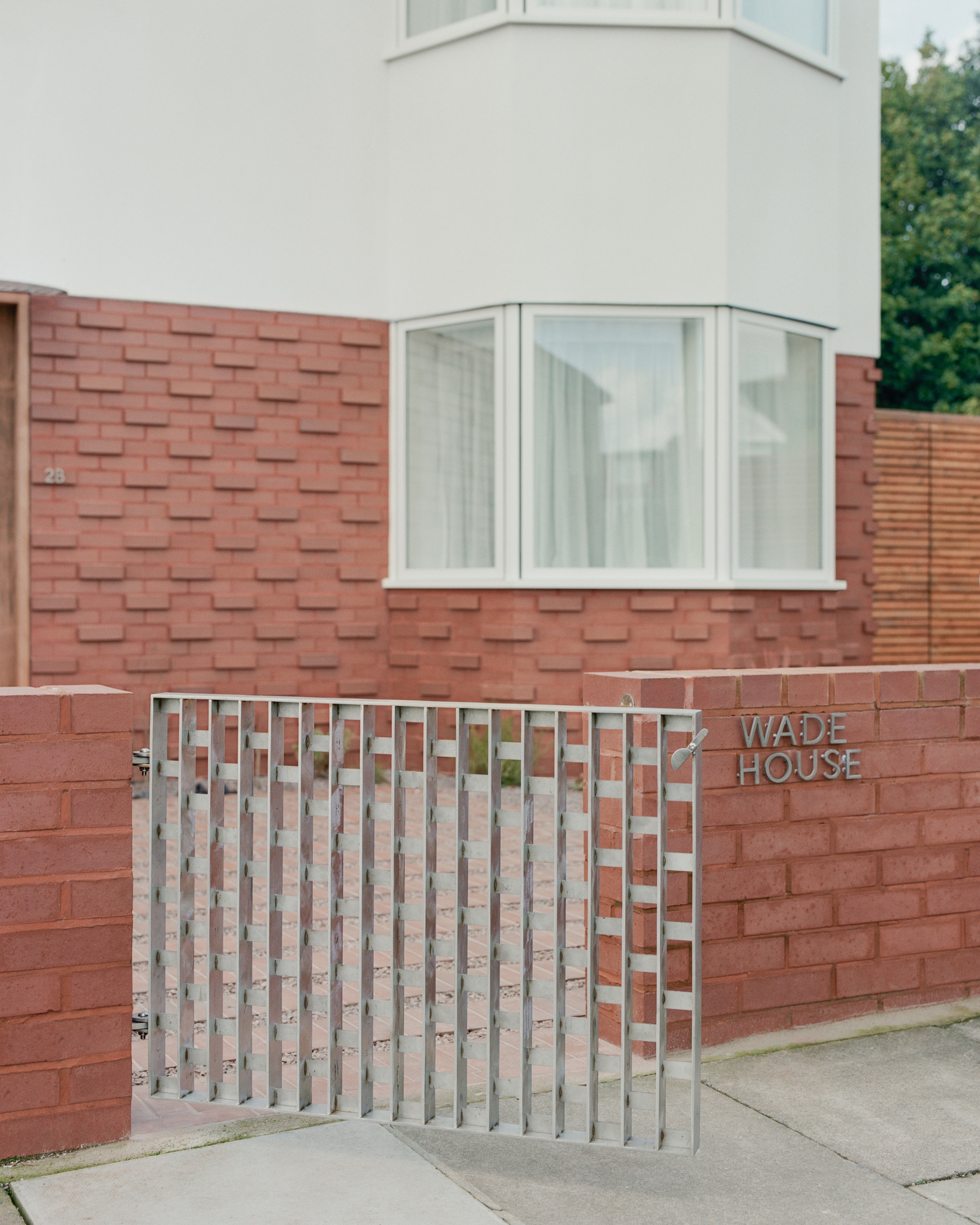 Red brick street wall with a slatted metal gate and a white bay window above, under overcast daylight