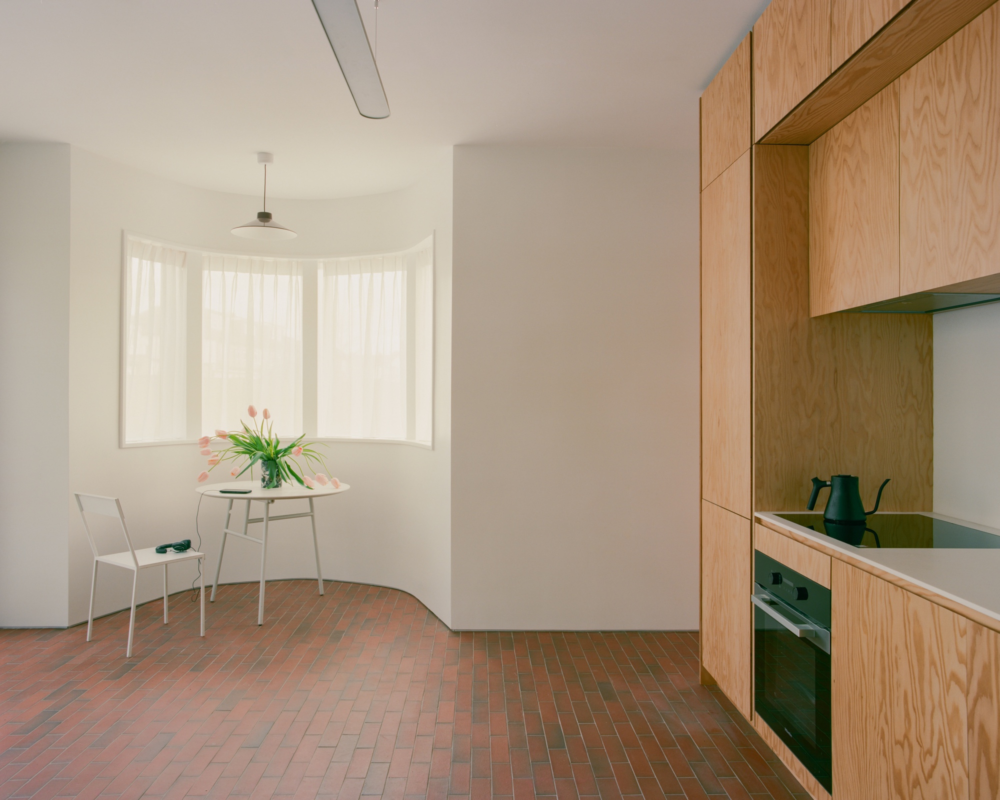 Curved bay window nook with a round table and tulips beside plywood kitchen joinery, on a red brick tile floor