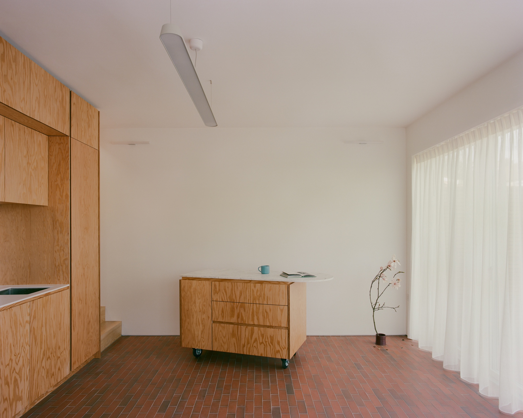 Wheeled plywood island with a stone top stands on a brick floor beside a floor-length sheer curtain in soft daylight