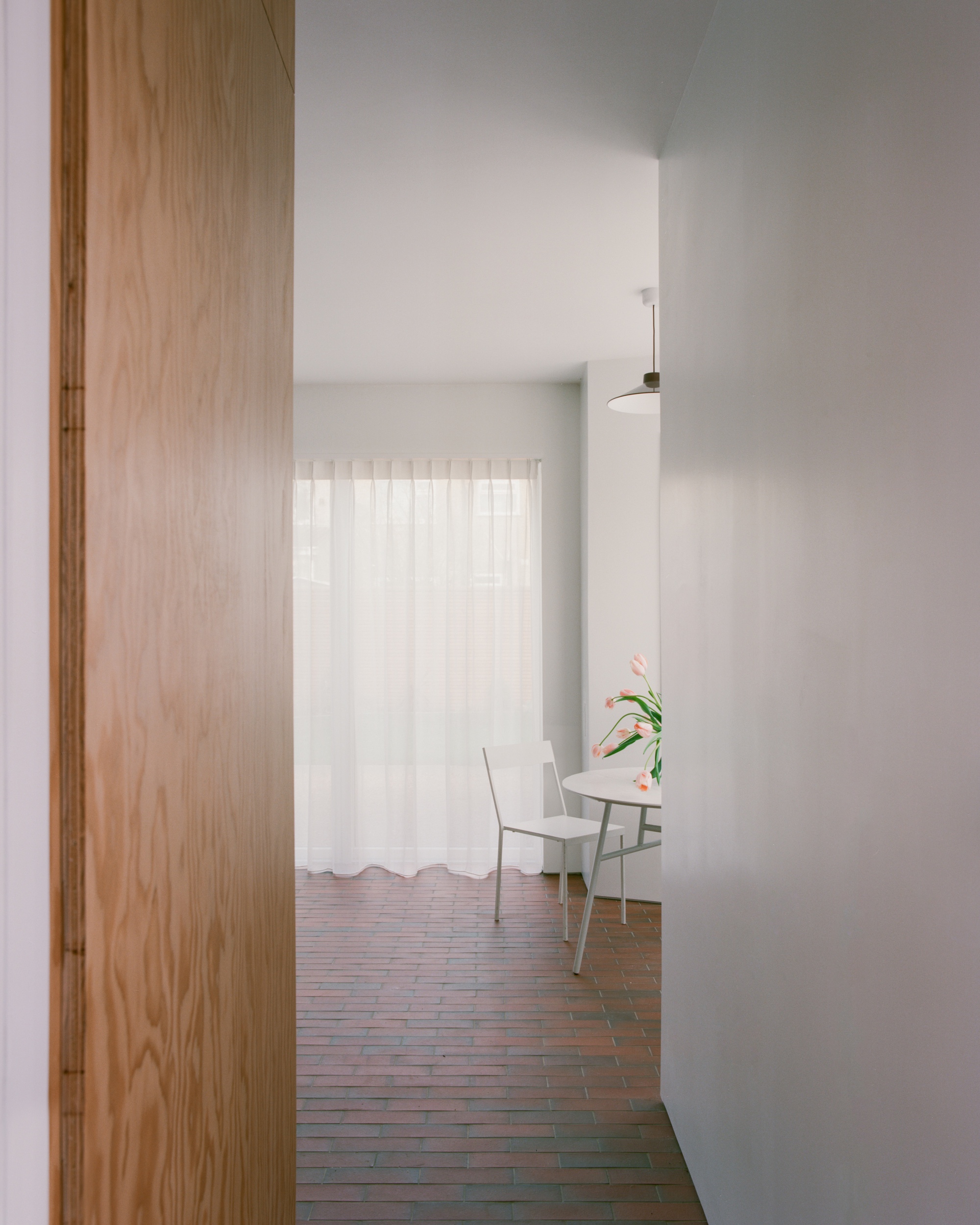 View past a plywood panel edge toward a dining chair and tulips in front of a sheer curtain