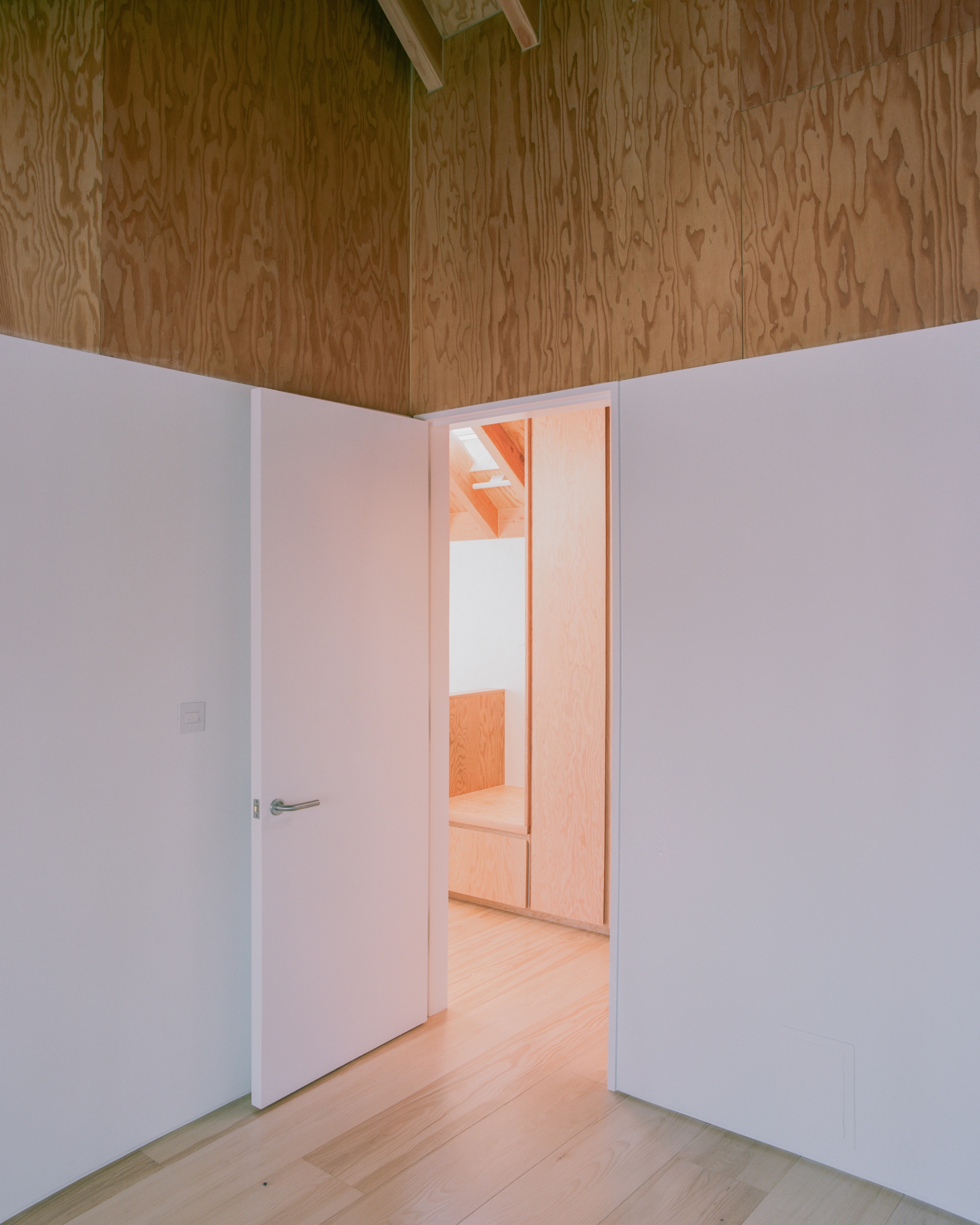 Open white door in a plywood-clad room leading to a sunlit space with built-in joinery and pale timber floor