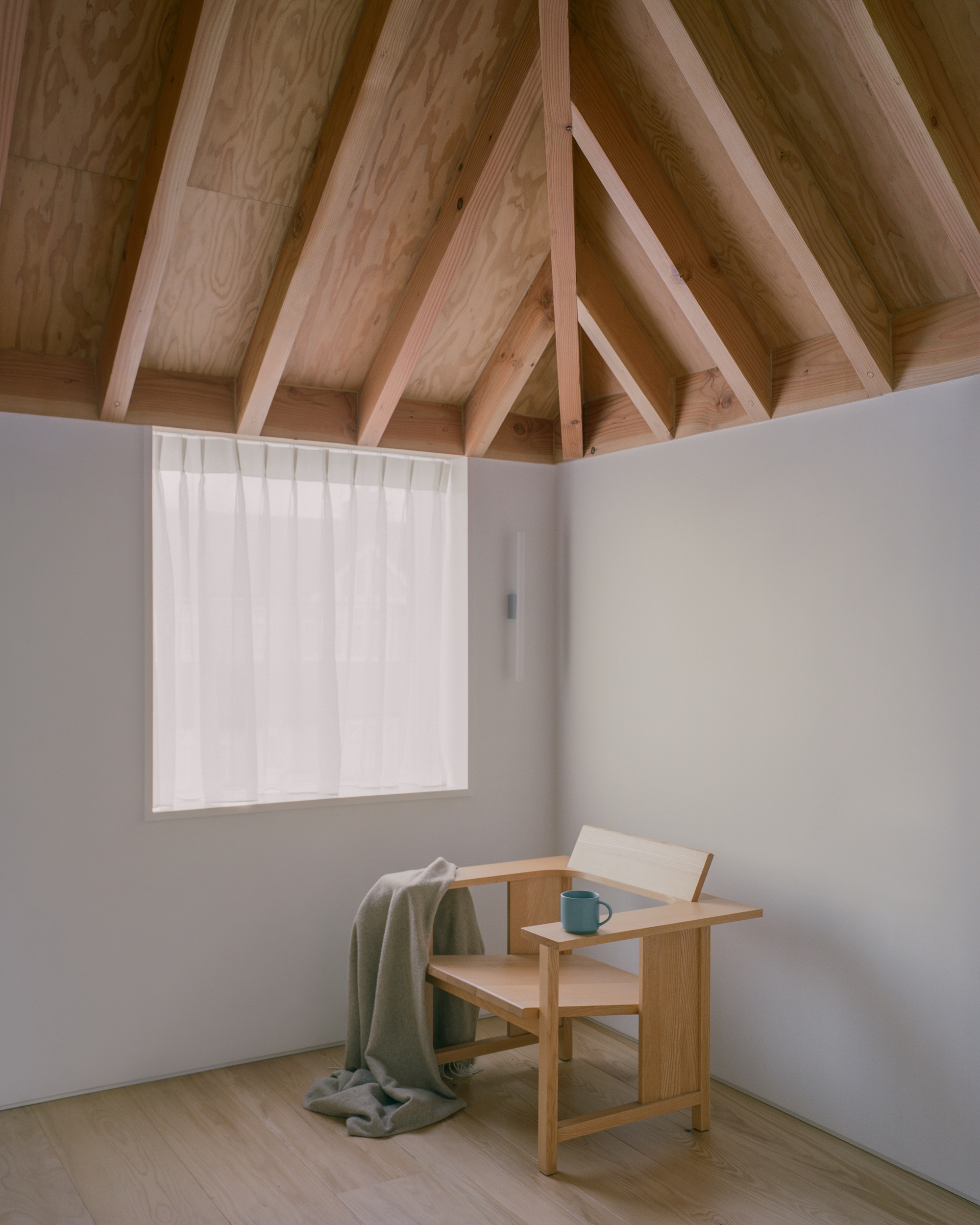 Timber hip roof rafters above a corner with a wooden armchair, grey blanket and blue mug beside a curtained window