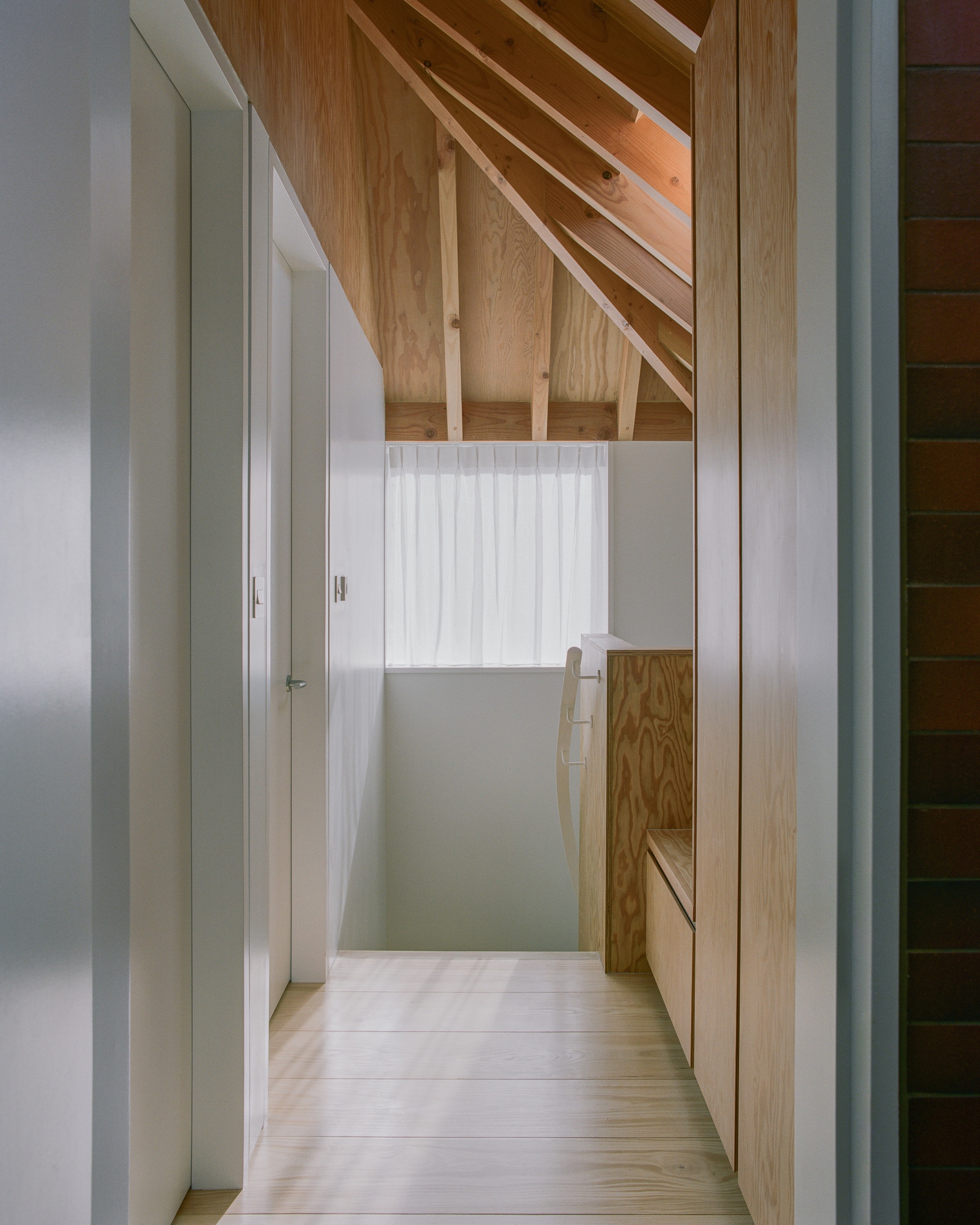 Upper landing with a pitched plywood ceiling, sheer curtained window and pale timber floor beside a red brick wall