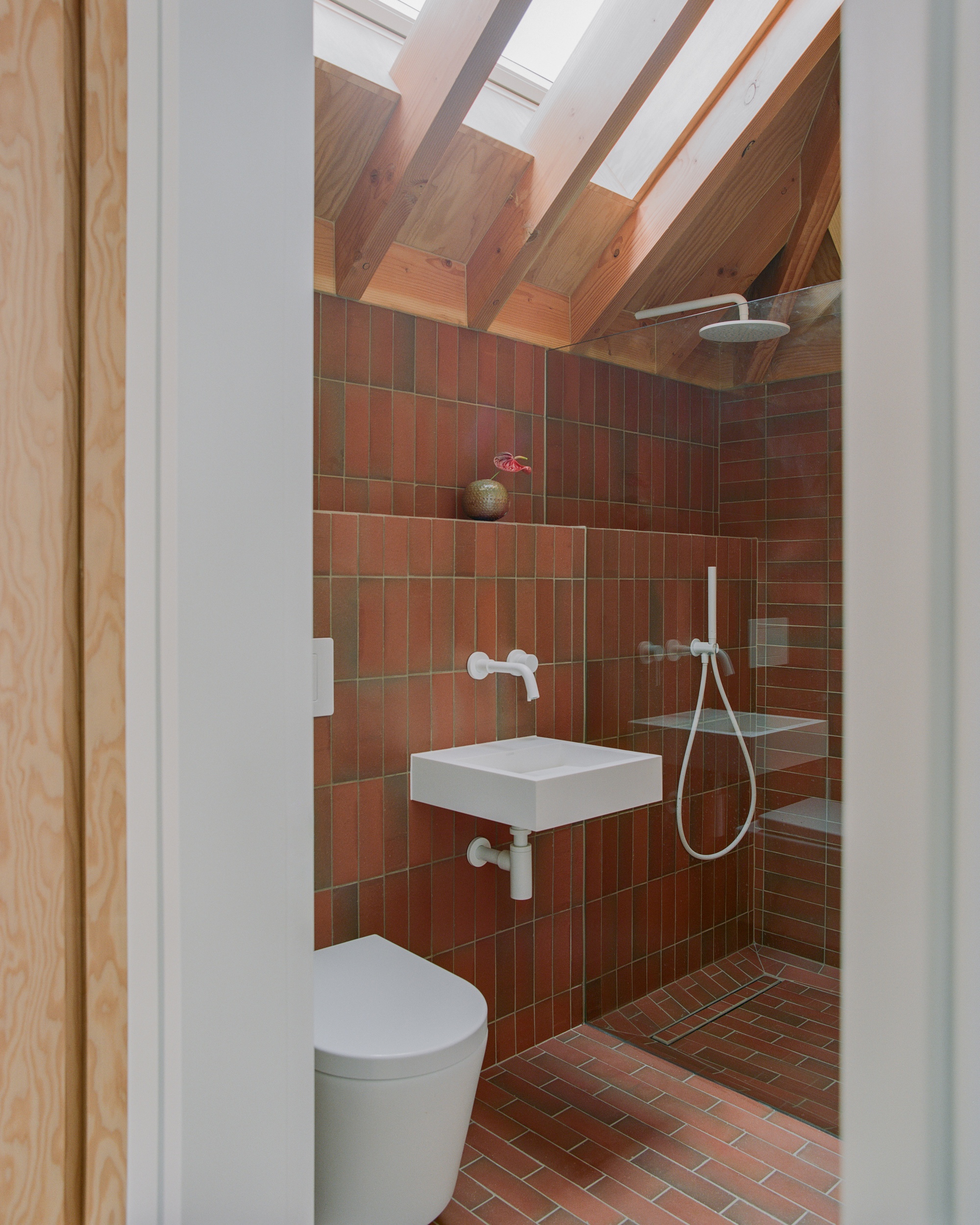 Compact bathroom with red brick-pattern tiles, white wall-hung basin and rain shower beneath rafters and a skylight