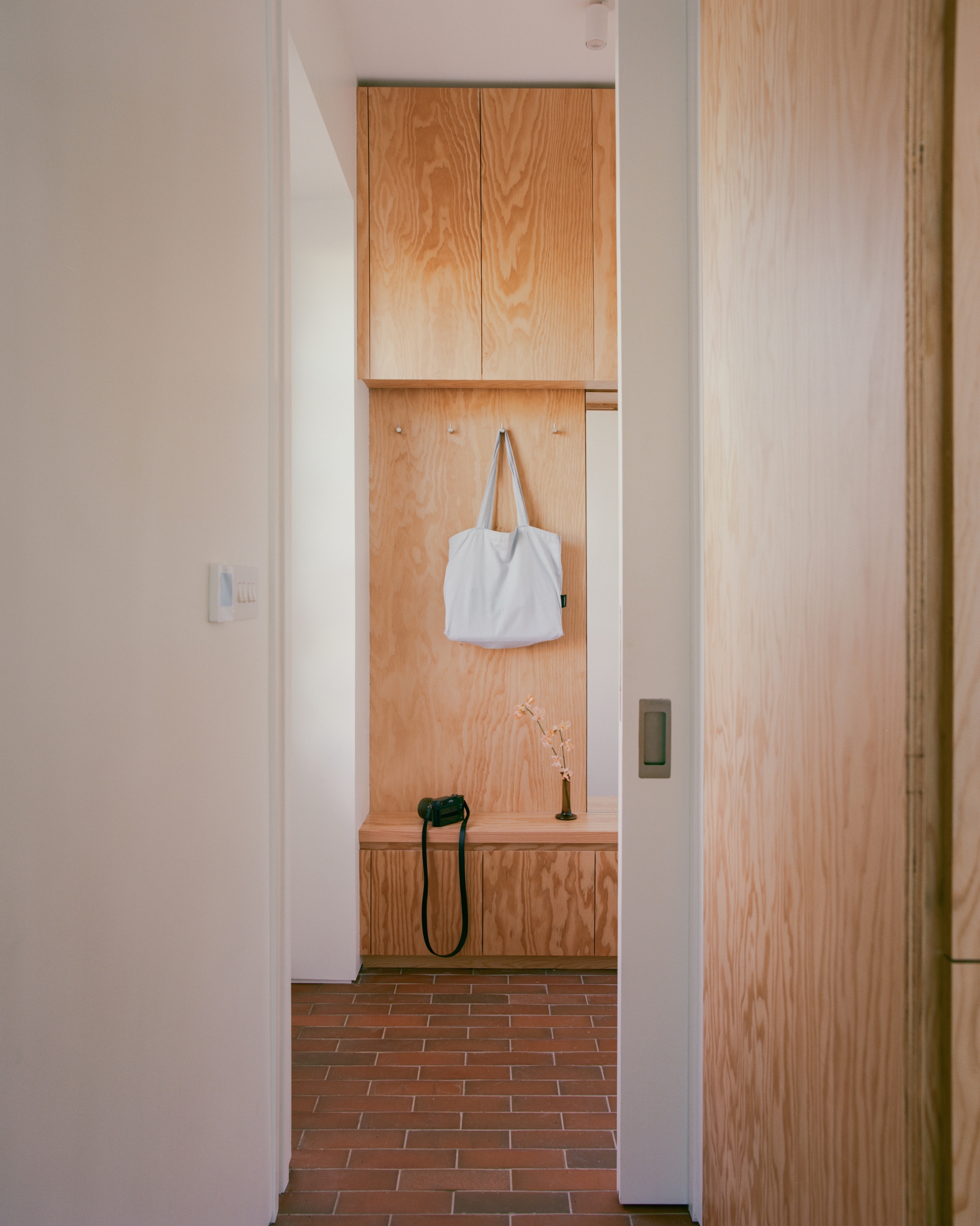 Entry nook seen through a sliding door, with plywood cabinets, a canvas bag on a hook and a camera on the bench