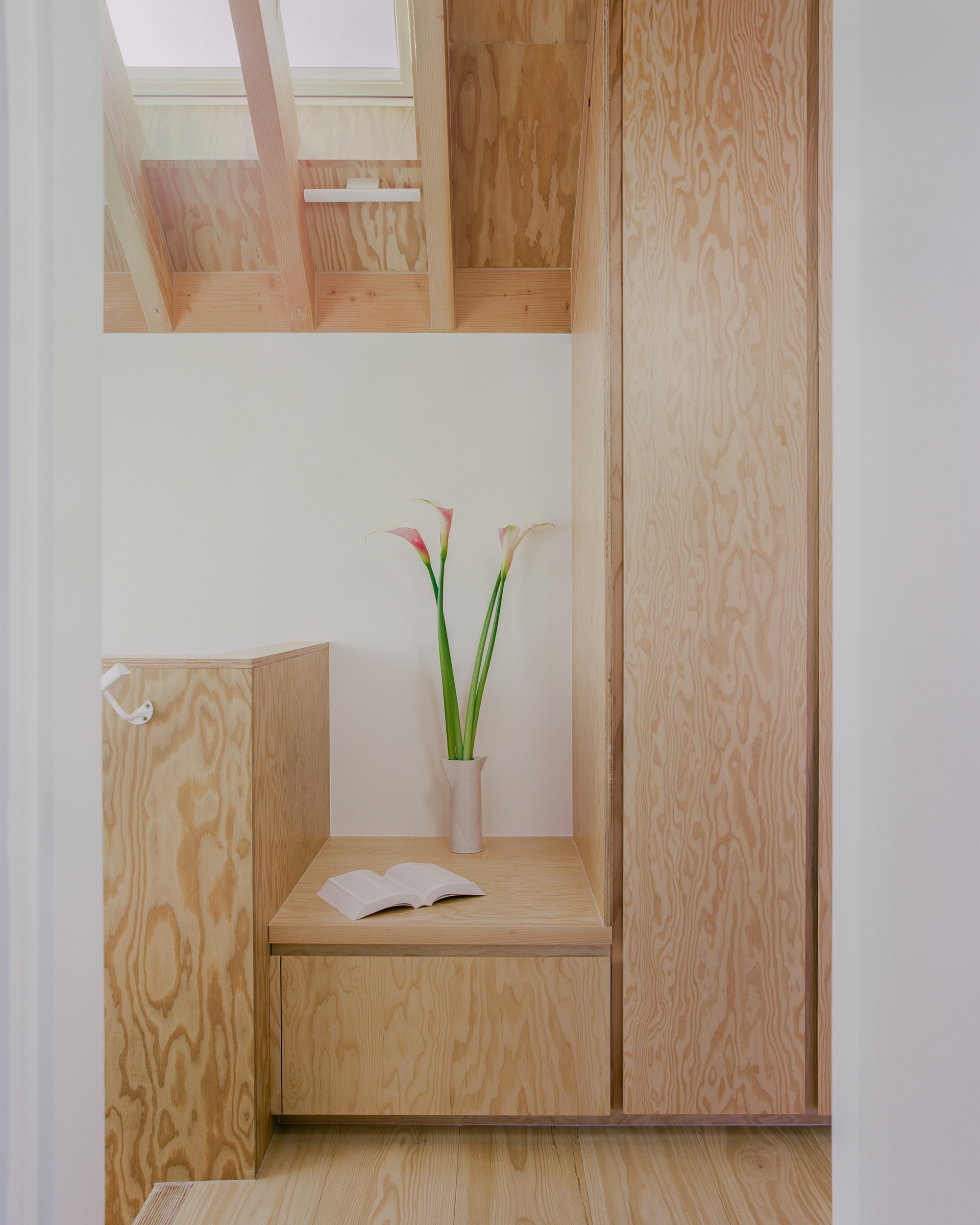 Plywood built-in bench and tall cupboard on a landing, with calla lilies in a vase and an open book in soft light