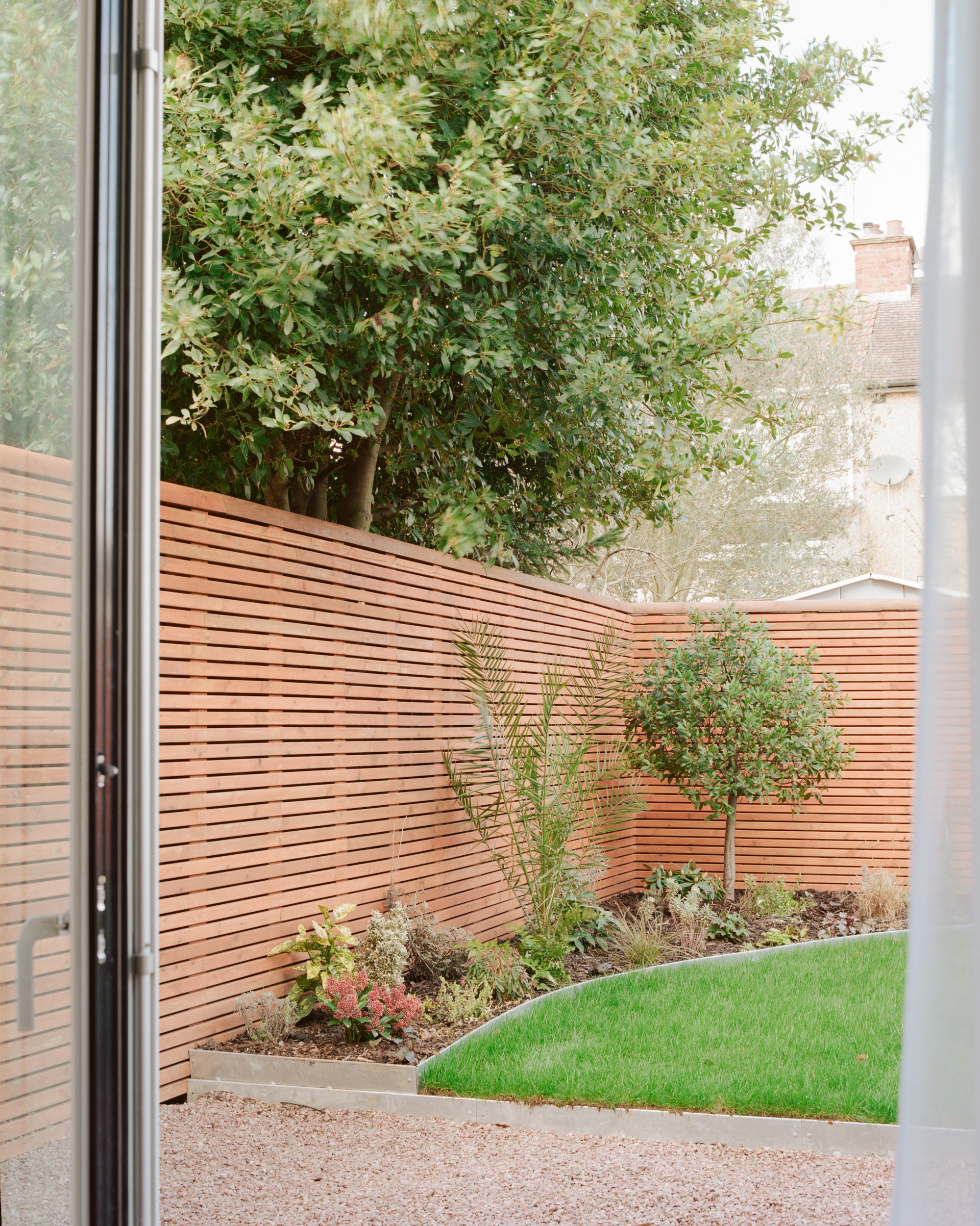 View through glass doors to a slatted timber fence, lawn, planted border and a small tree on an overcast day