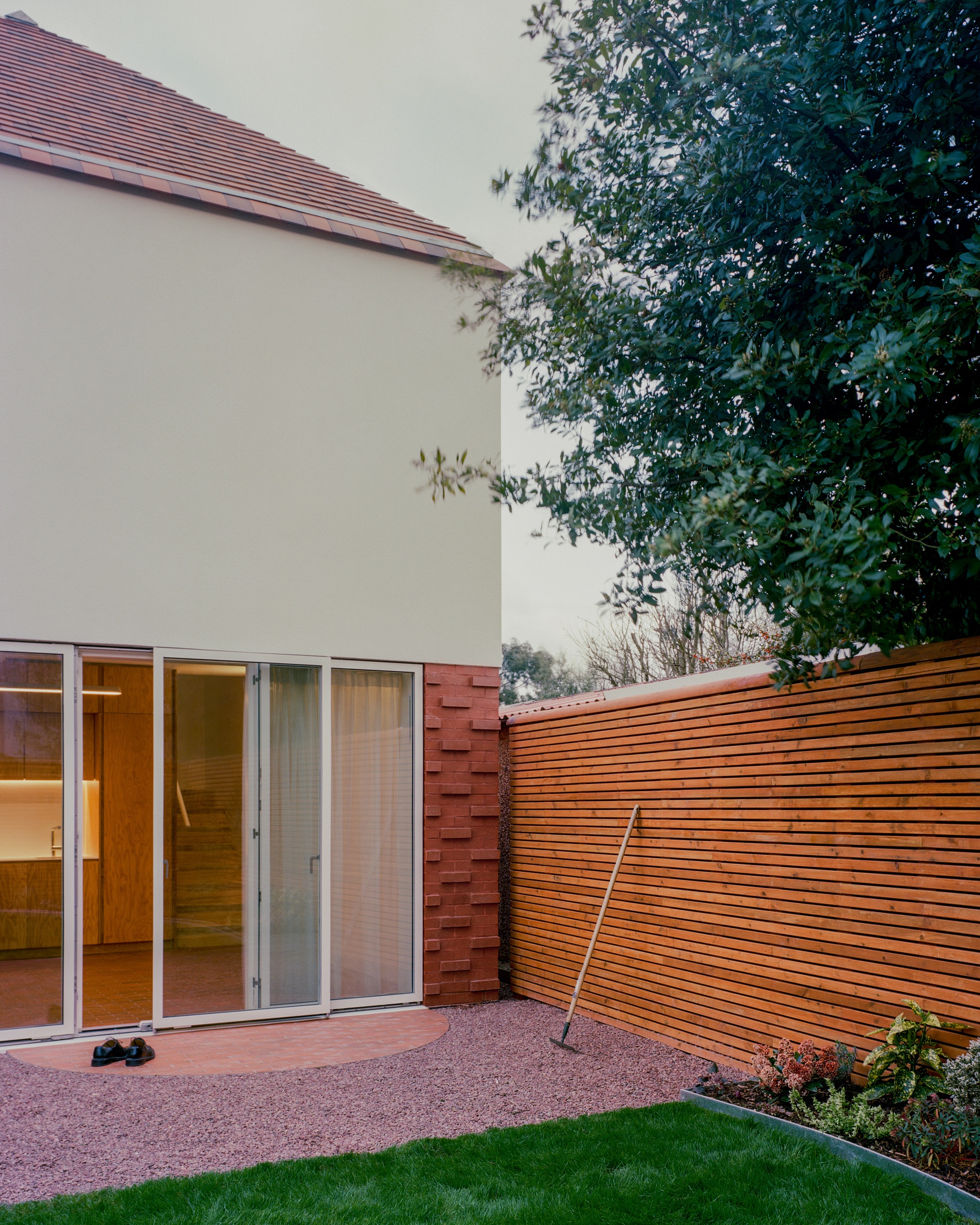 Rear corner with sliding glazed doors, a patterned red brick edge and a timber slat fence, with a rake leaning against it at dusk