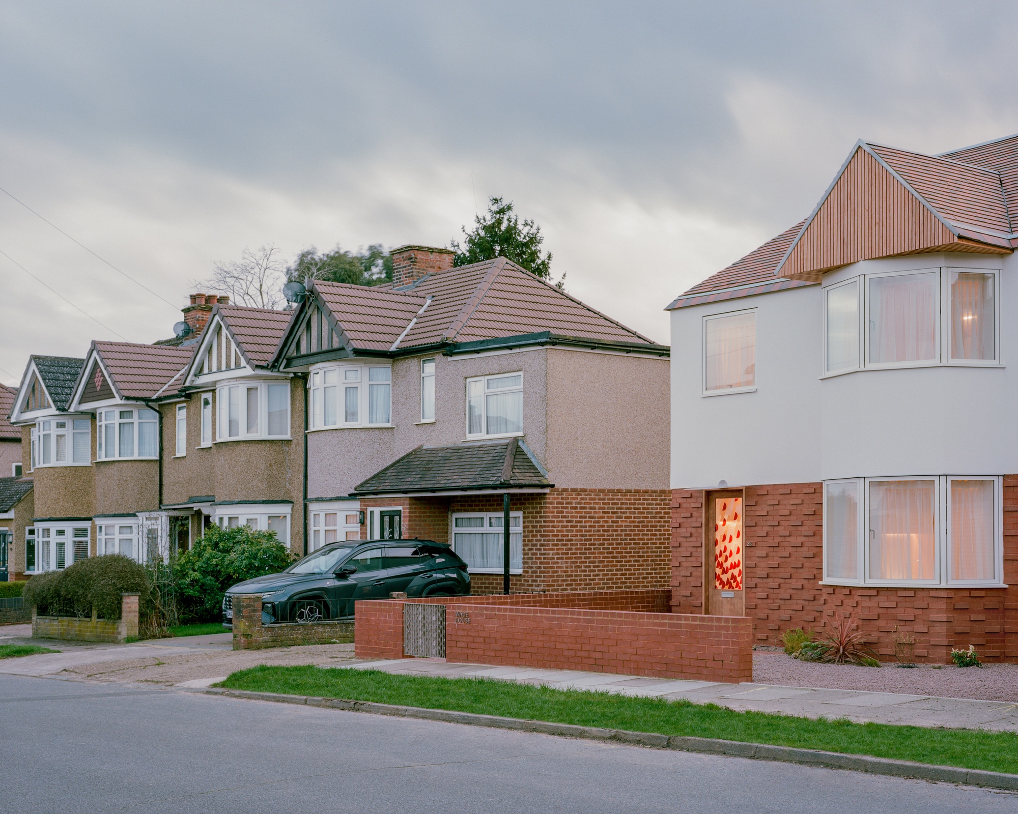 New house with brick base and timber gable beside a row of pebbledash semi-detached homes, a parked car in front at dusk