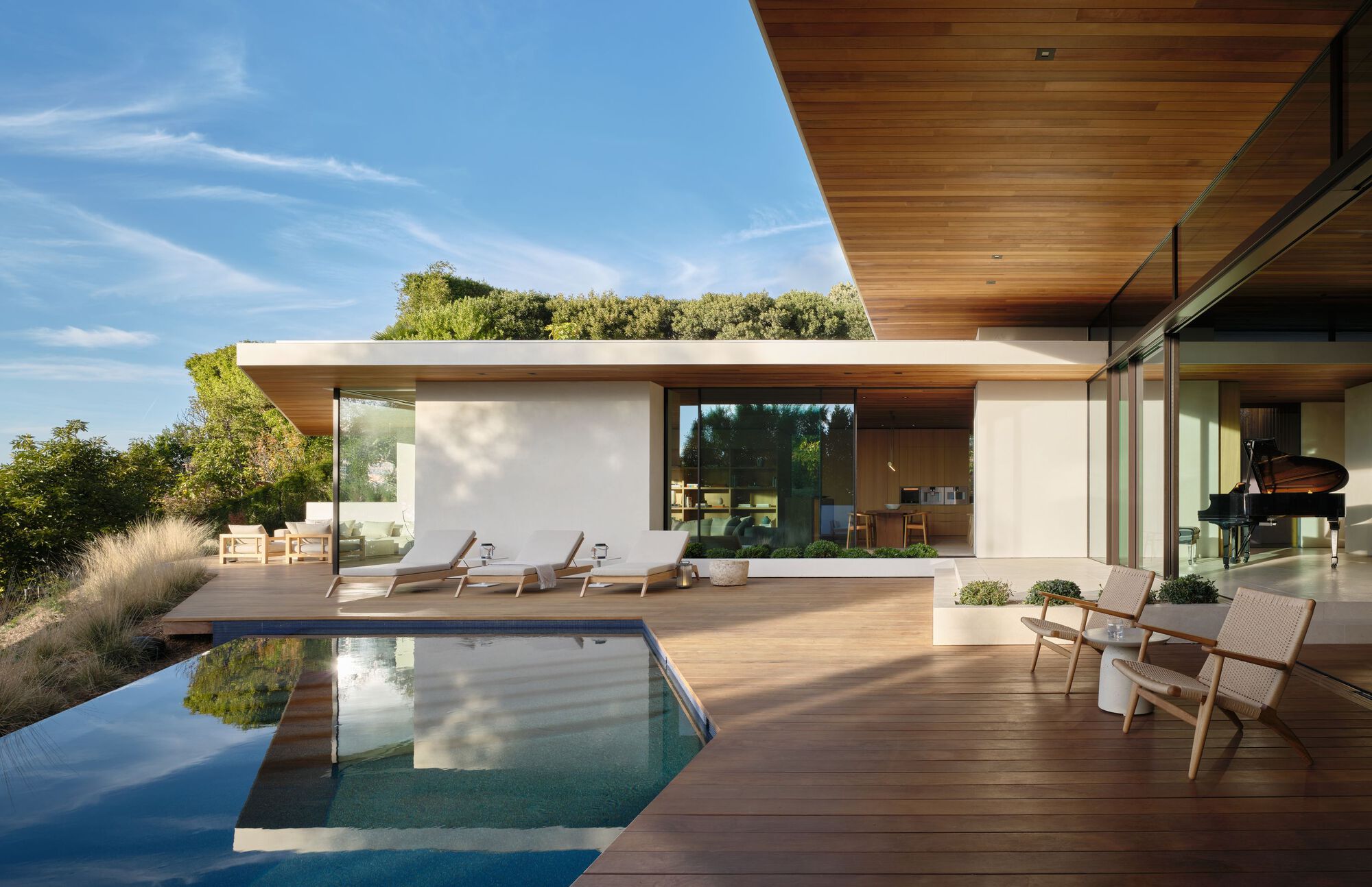 Timber deck with infinity pool and lounge chairs beside a glass-walled pavilion, a grand piano visible inside under a blue sky
