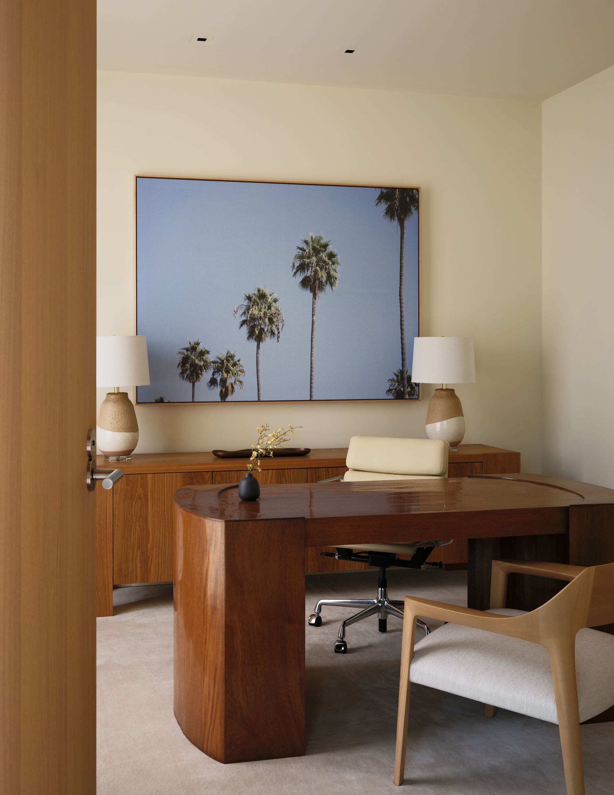 Study with a curved walnut desk, cream leather chair and a framed palm tree photograph flanked by two table lamps