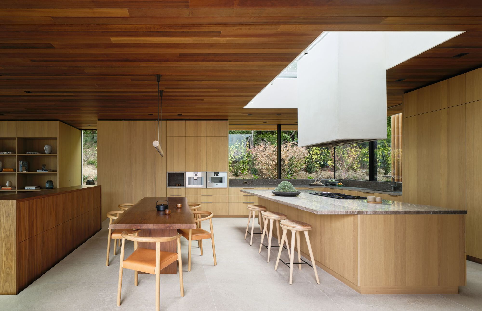 Kitchen with oak cabinetry, stone-topped island, bar stools and a white skylight shaft above, garden glimpsed through the window band