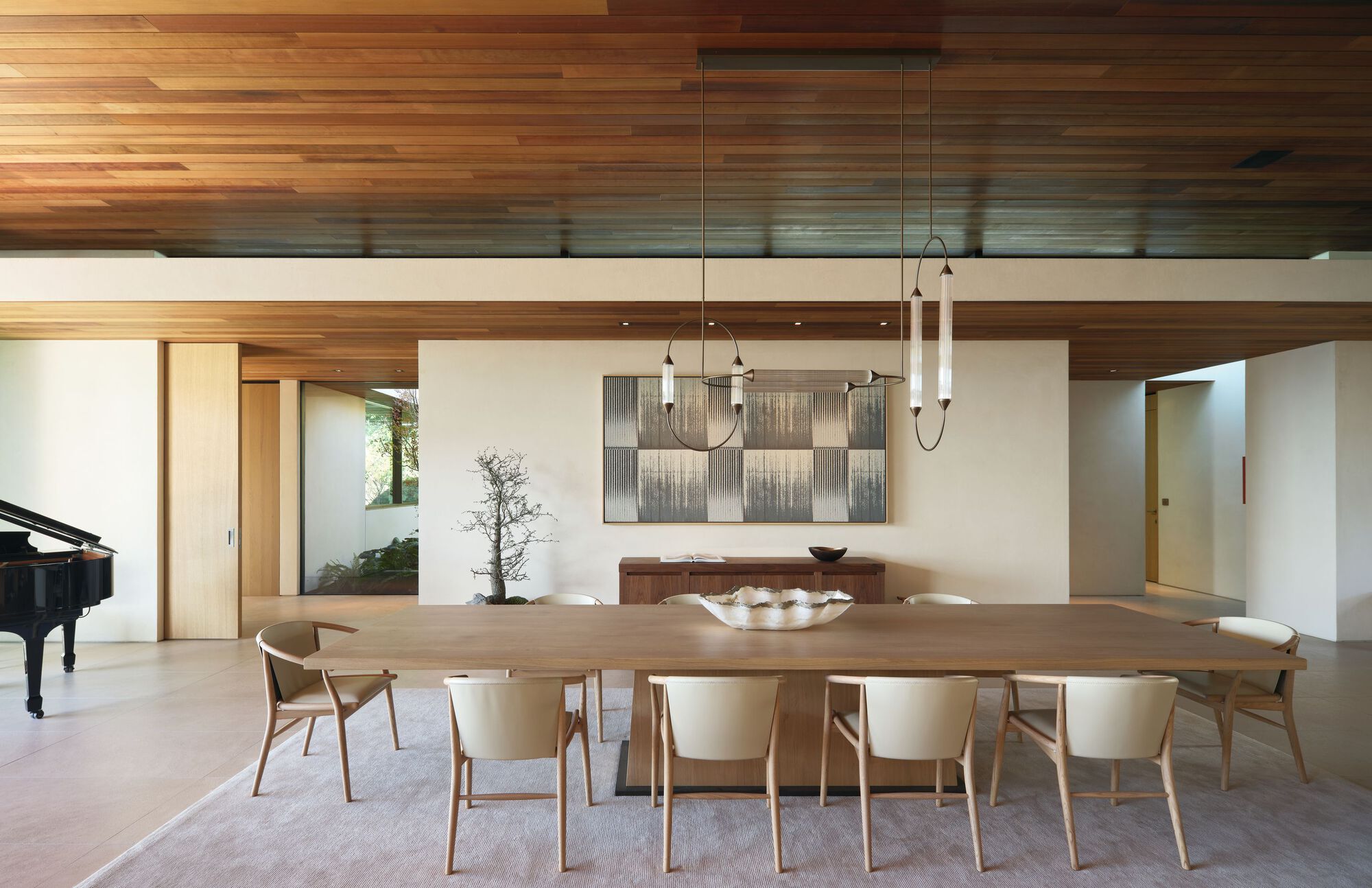 Long oak dining table with cream chairs and two pendant lights under a clerestory and timber ceiling, a piano at left