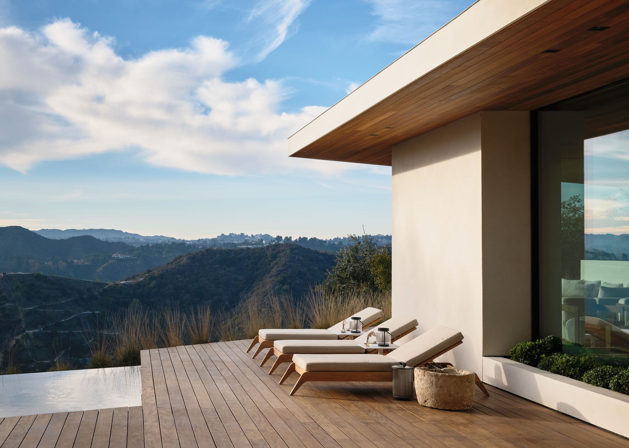 Timber deck with four sun loungers beside an infinity pool edge, overlooking hazy hills under a cloud-streaked sky