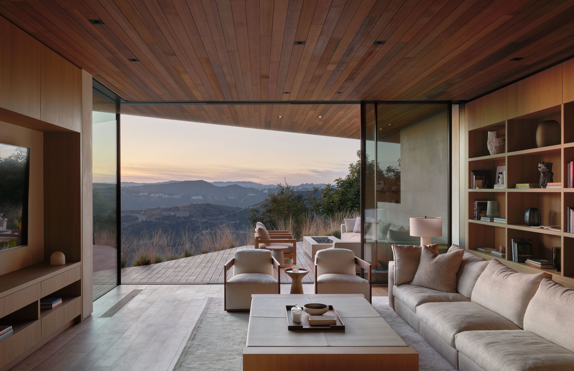 Sitting room opening through corner glazing to a deck and layered hills at dusk, with built-in shelves and a sectional sofa
