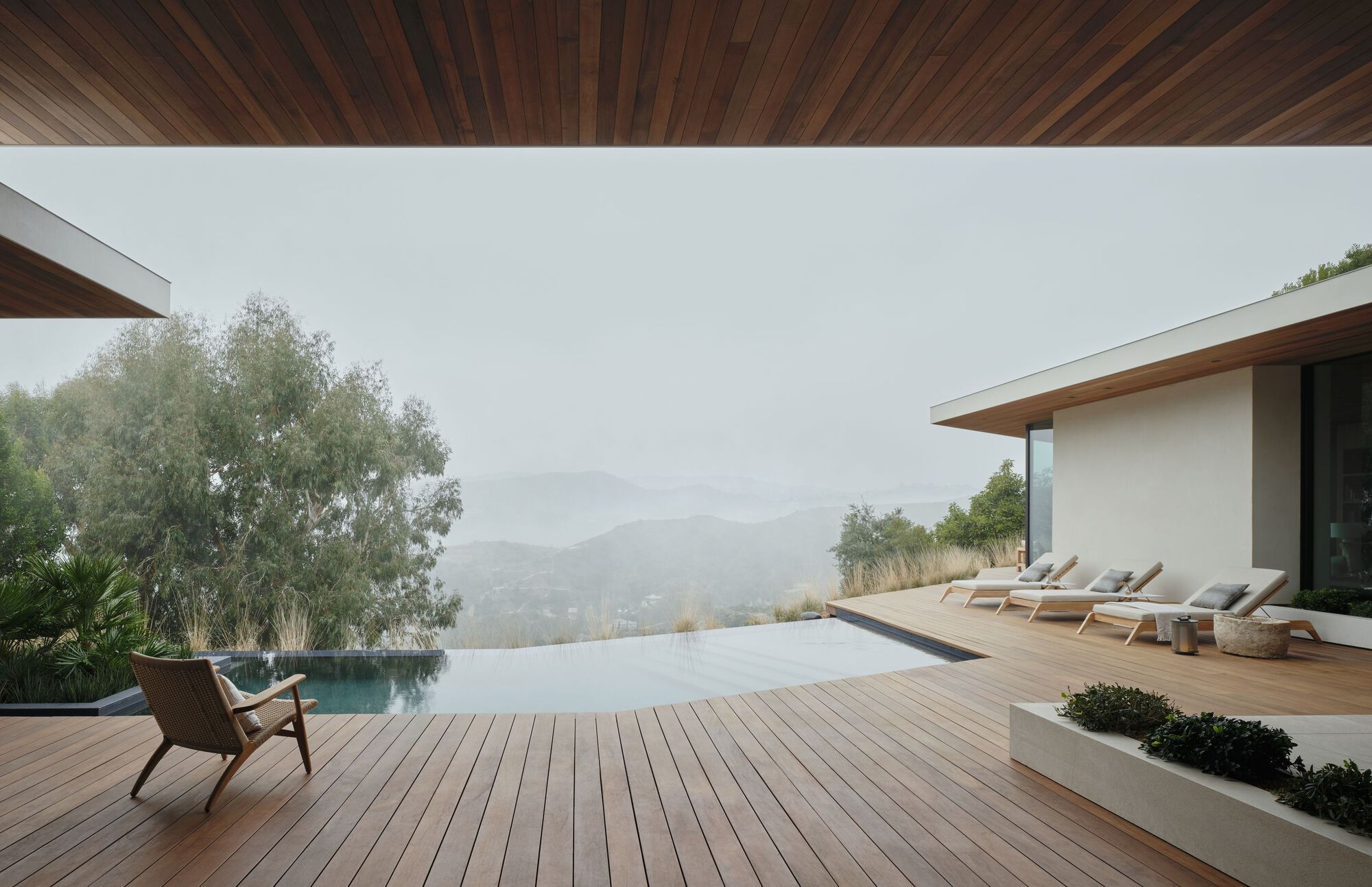 Timber deck with an infinity pool and lounge chairs under a deep wood-lined overhang in heavy fog