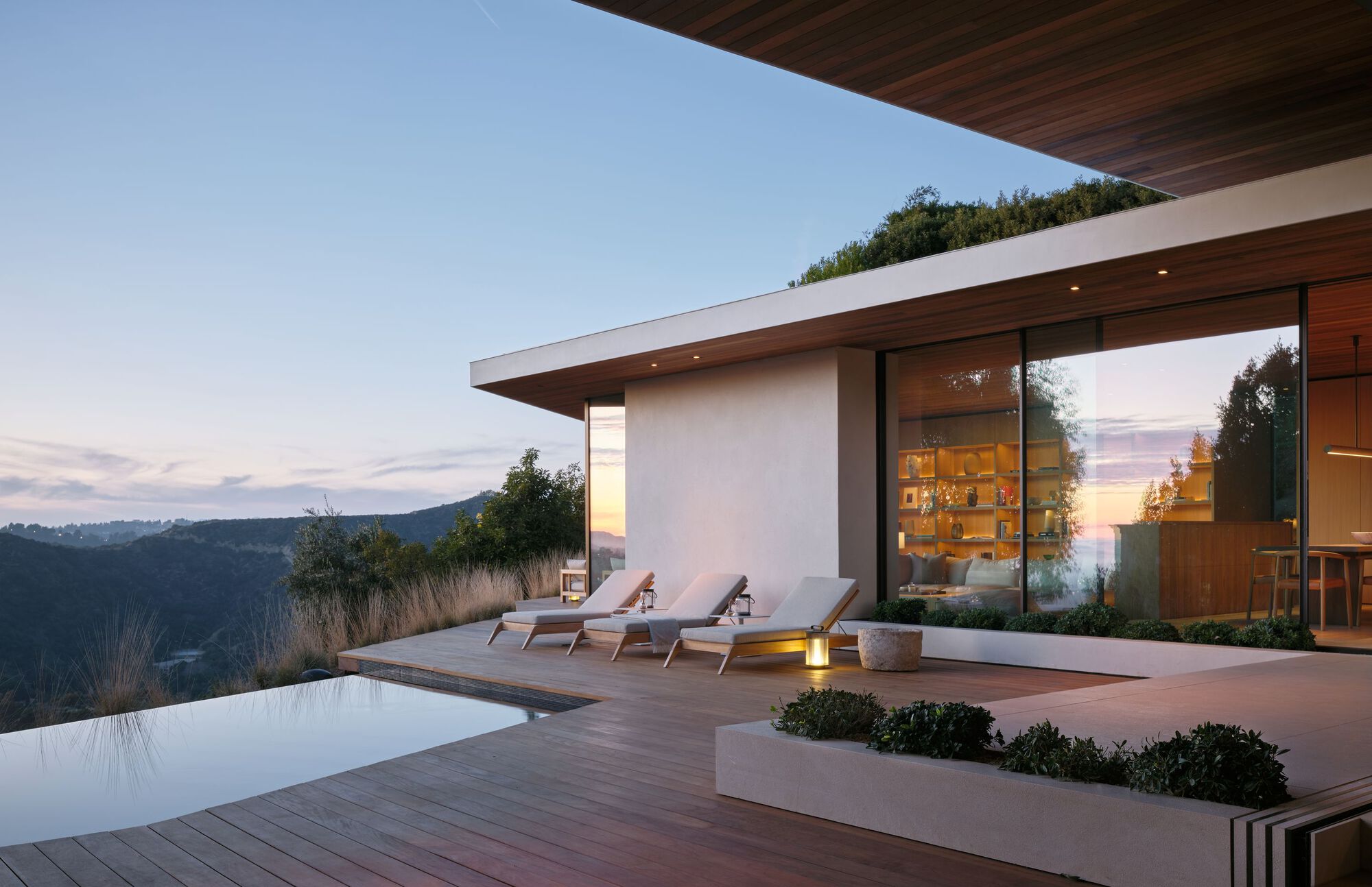 Pool terrace with three lounge chairs and a planter bed beside a glazed wall, glowing at dusk