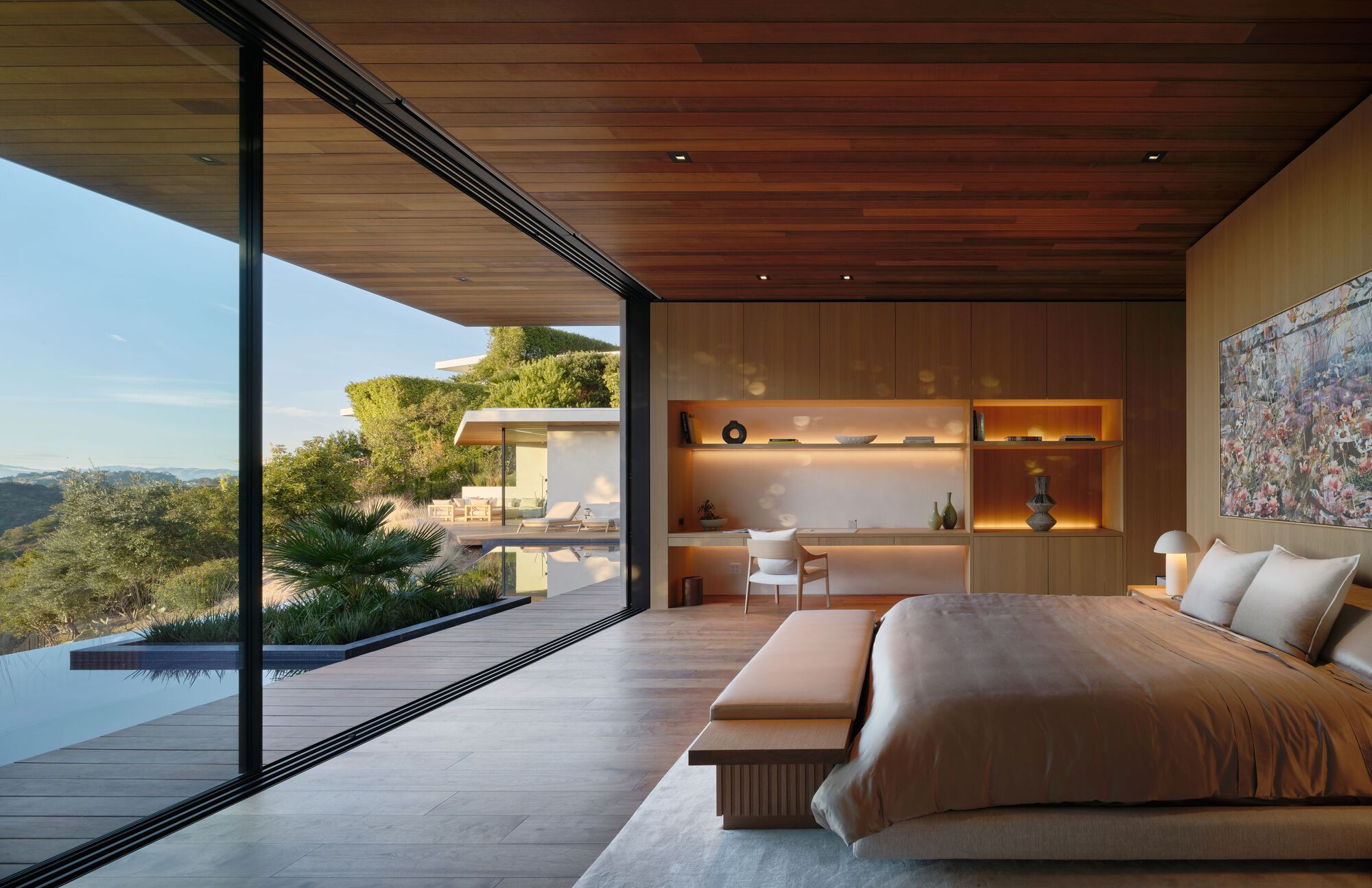 Bedroom with a wood ceiling and built-in shelving, its glass wall slid open to the pool deck in late sun