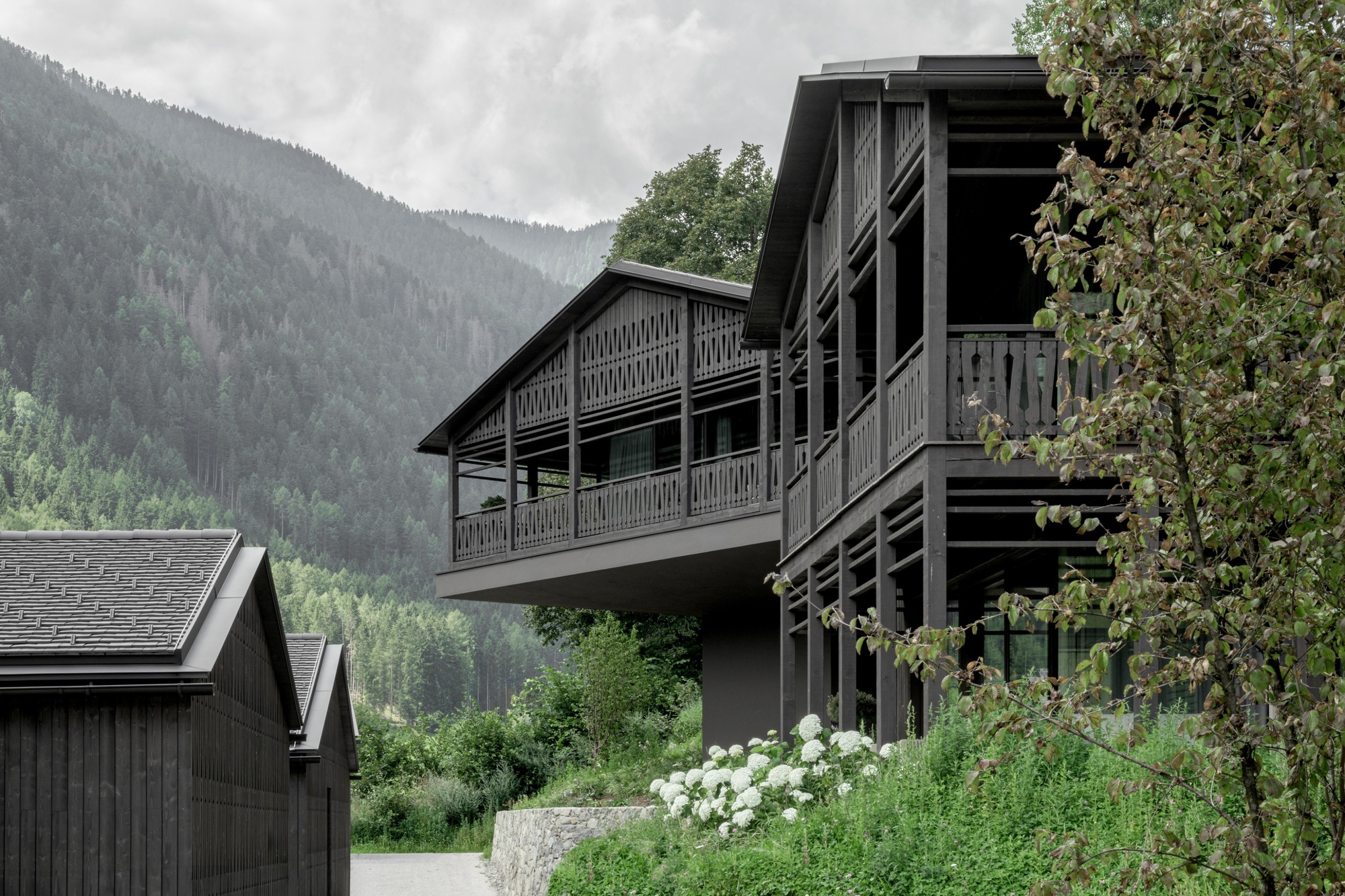 Cantilevered upper floor with timber balustrades and columns beside a slate-roofed outbuilding, with white hydrangeas in the foreground