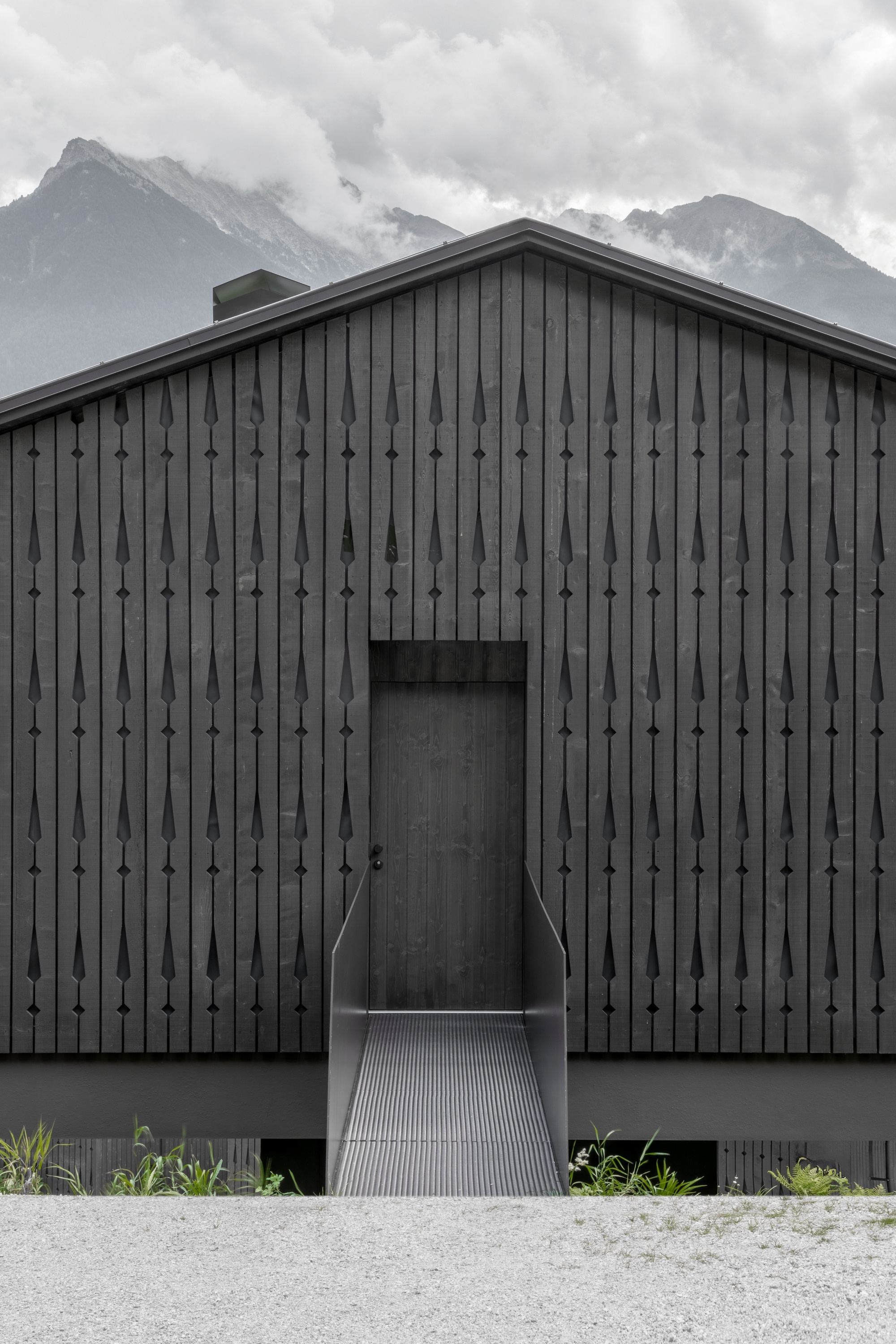 Gable facade of dark vertical boards with teardrop cutouts and a recessed door reached by a metal grate ramp, mountains behind