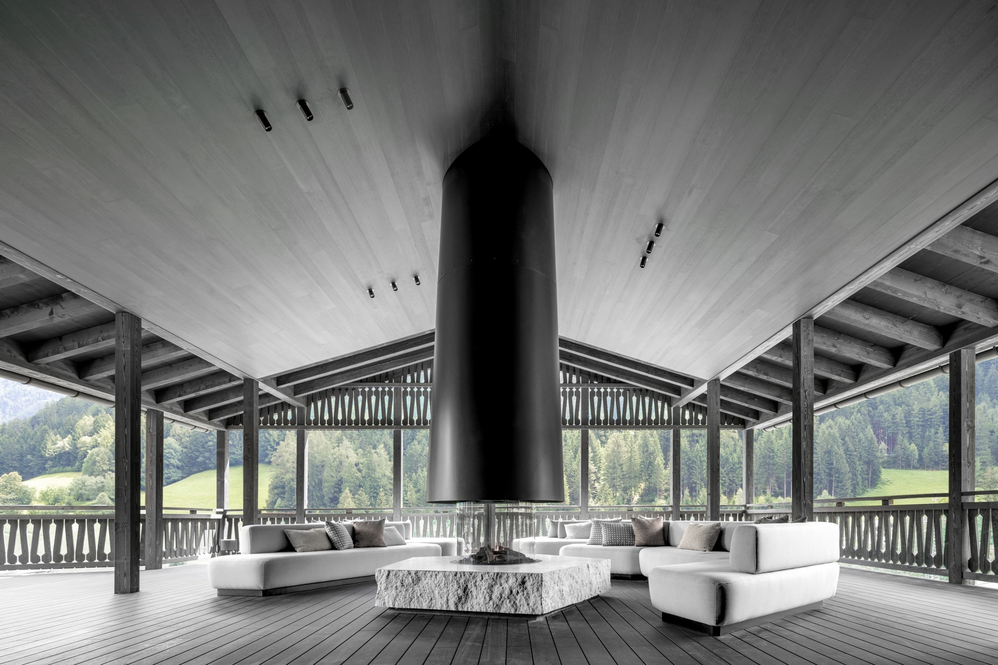 Open timber pavilion with a suspended black cylindrical fireplace over a stone block and white sofas, forested valley beyond the balustrade
