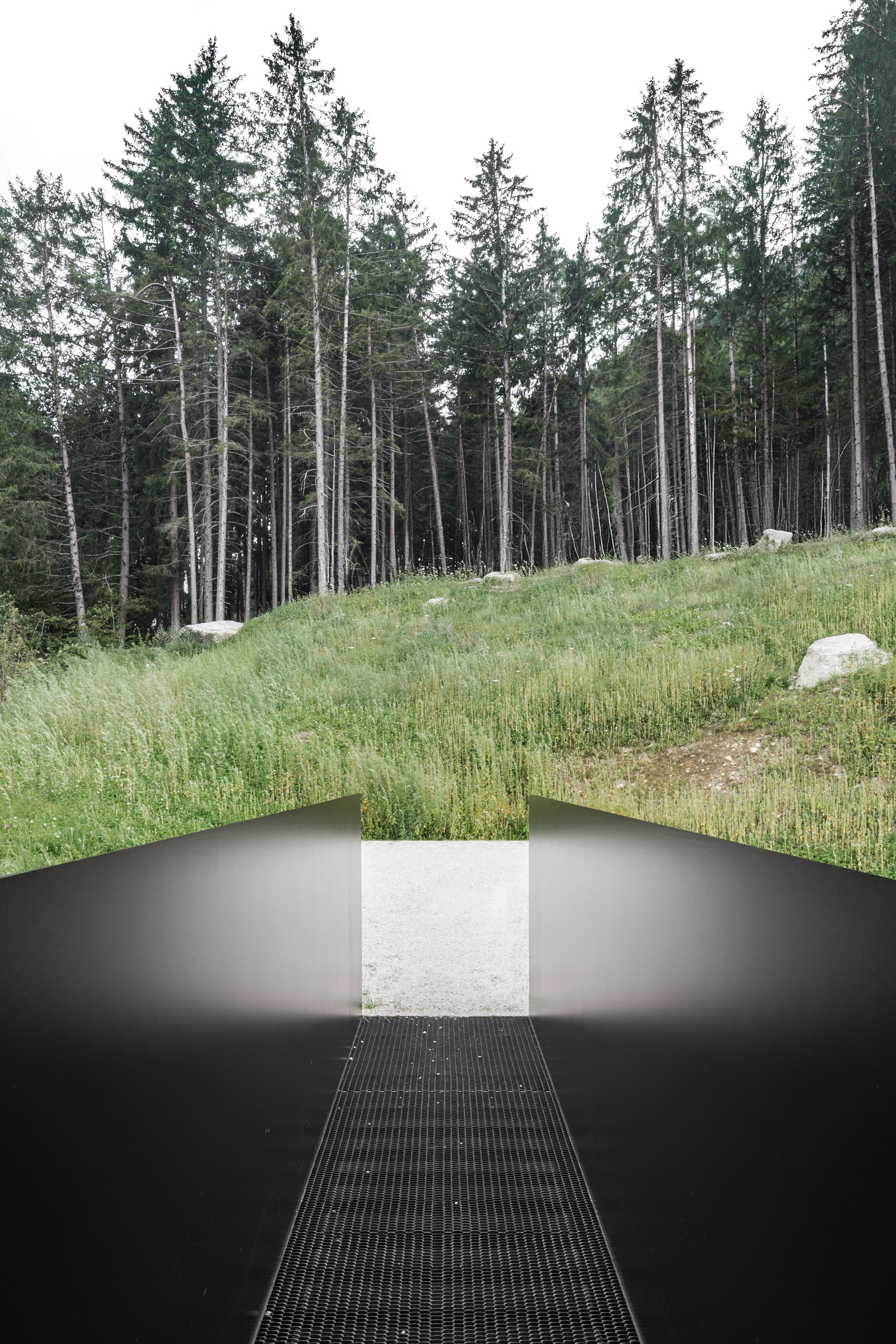 View along a steel grate walkway between dark angled walls toward a grassy slope and spruce trees on an overcast day