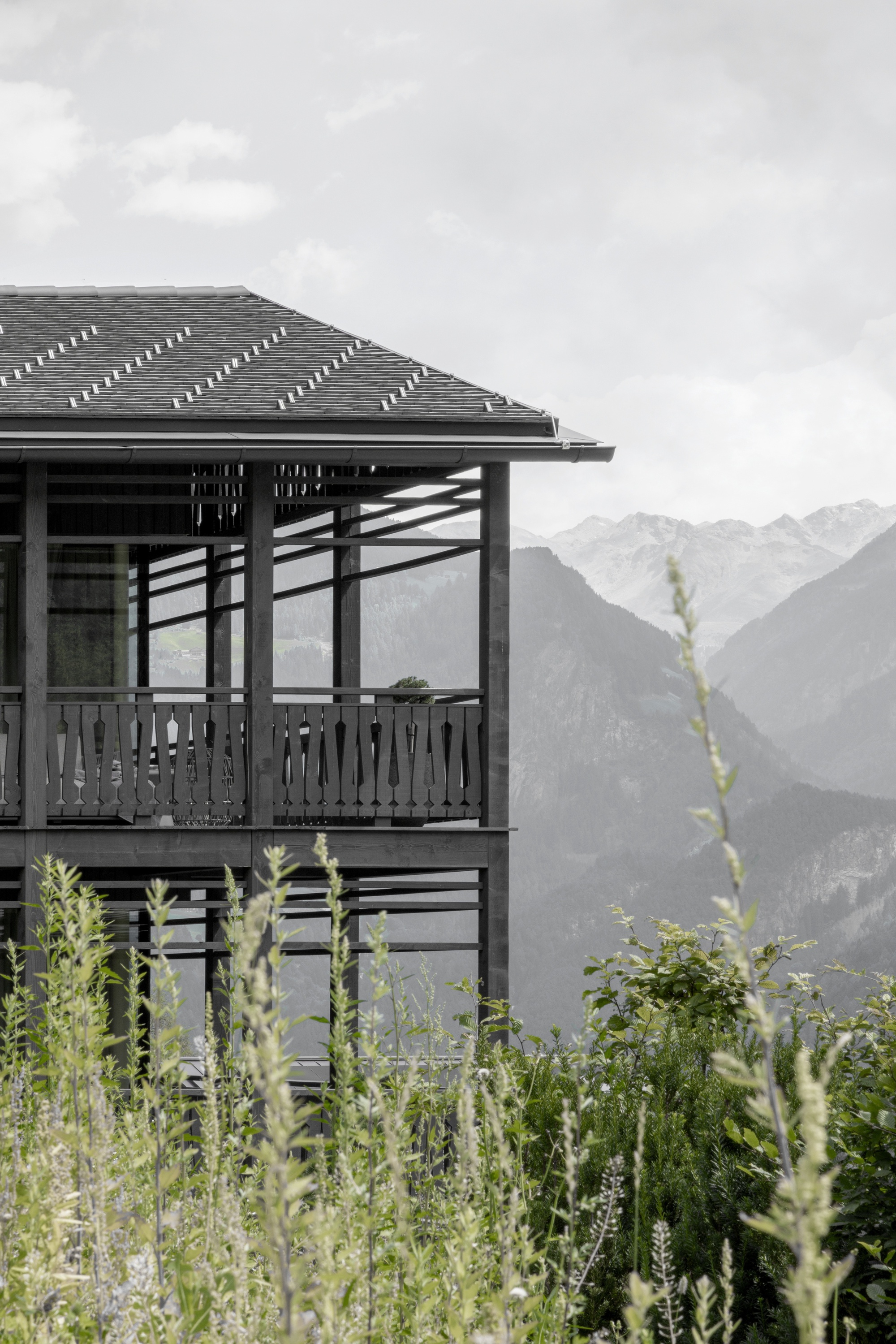 Timber chalet with a slate-grey roof and open balcony rising above tall grasses, with hazy mountain ridges behind under overcast sky