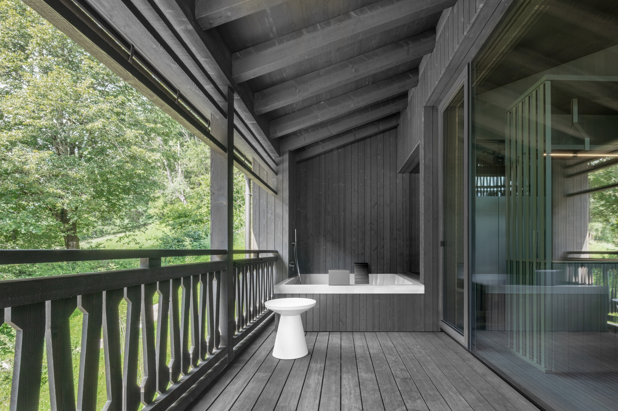 Covered timber balcony with a built-in bathtub, white stool and carved balustrade, looking out to green trees in summer light
