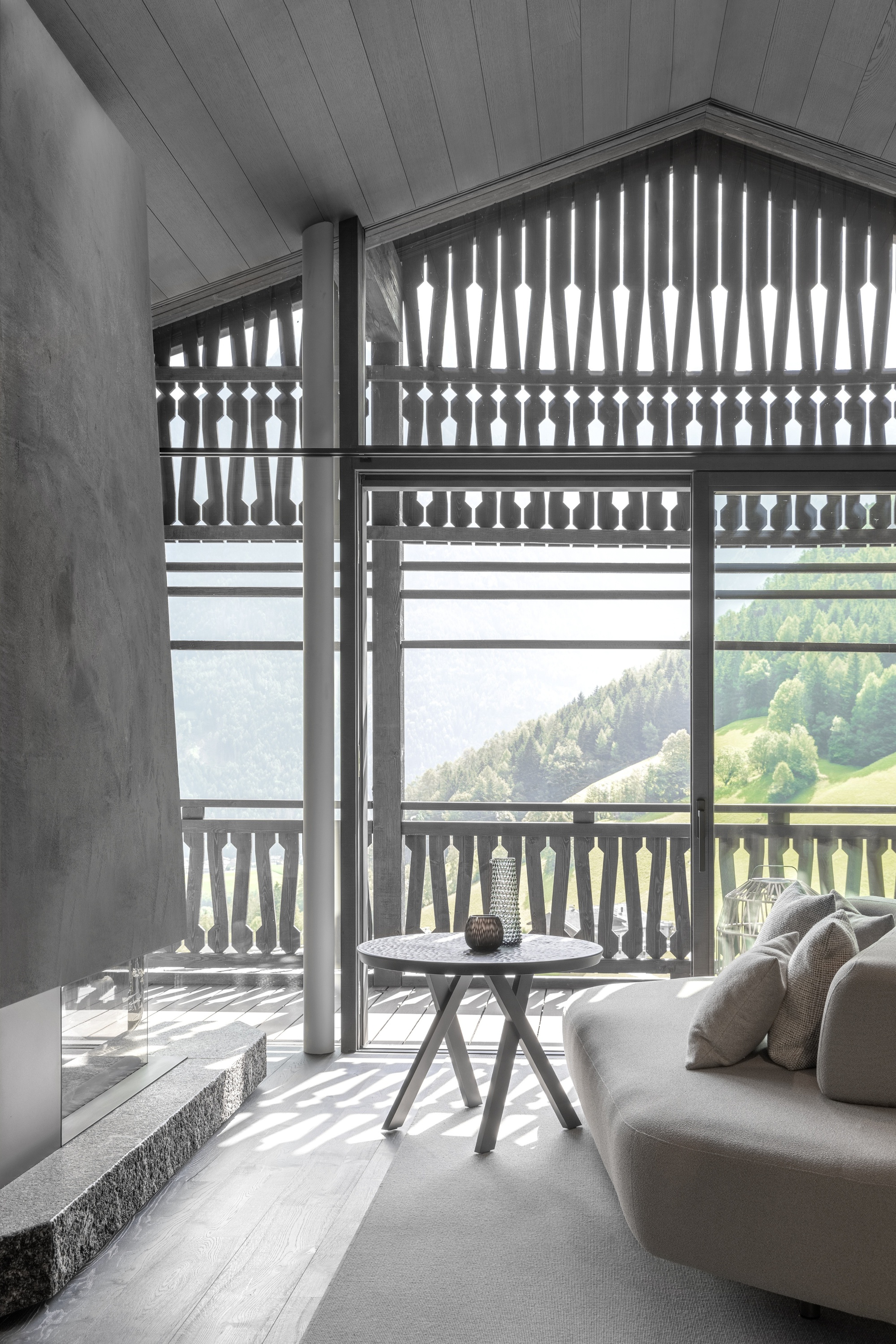 Living room corner with a curved sofa and round table, sunlight casting shadows through the fretwork screen toward a forested valley