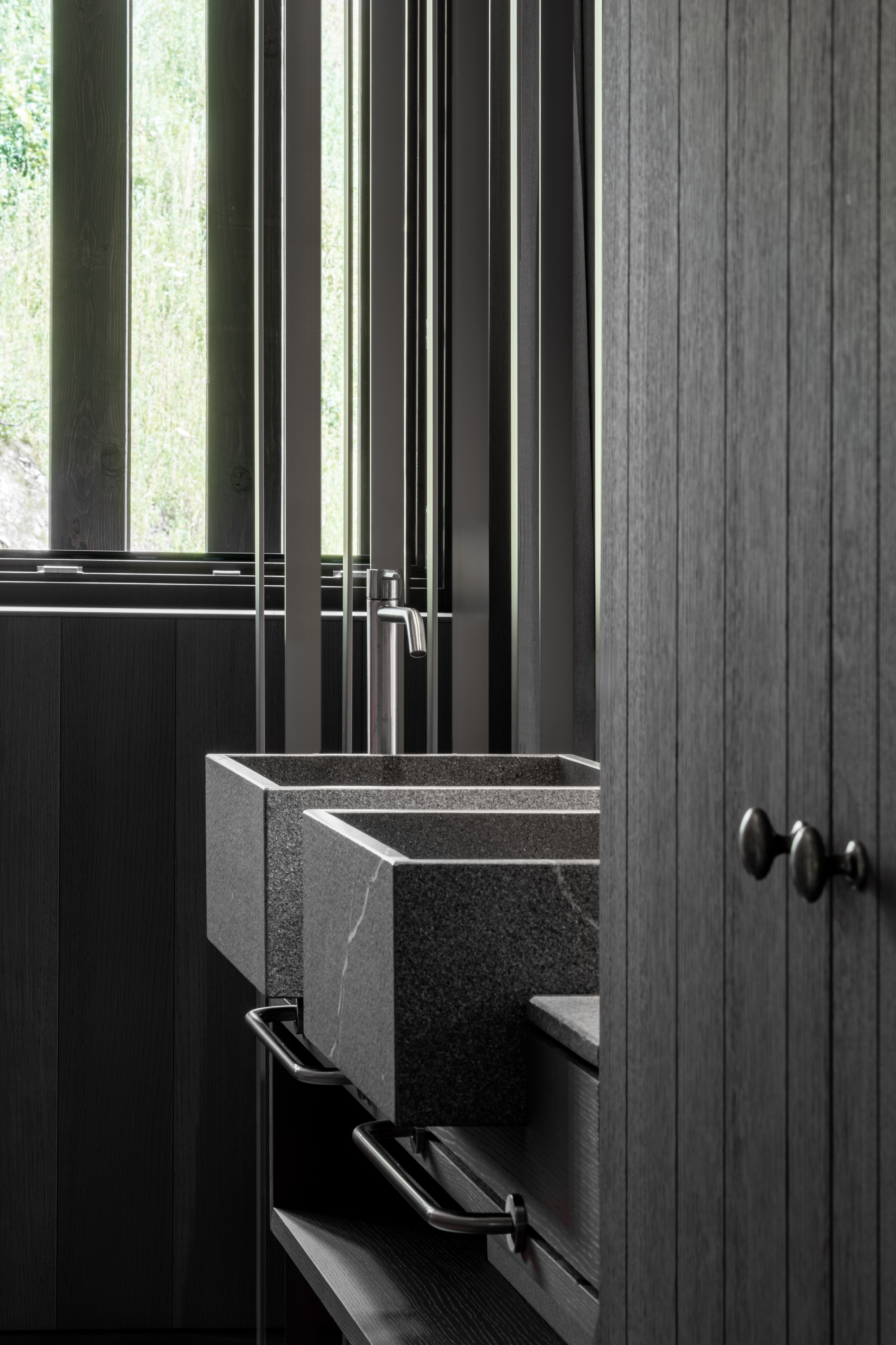 Close-up of two stone washbasins with a steel tap beside a dark vertical-board timber door and slatted window