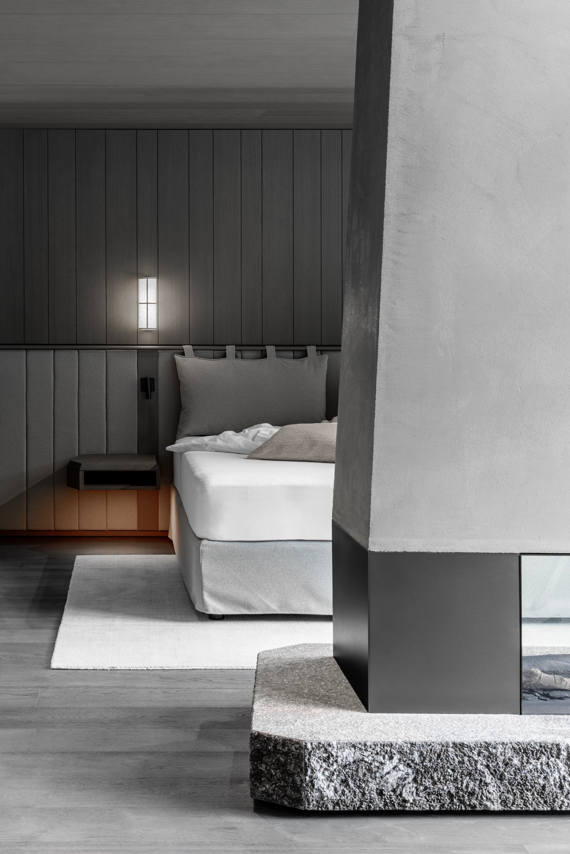 Bedroom with a panelled headboard wall and wall lamp, seen past a plastered fireplace block resting on a rough granite base